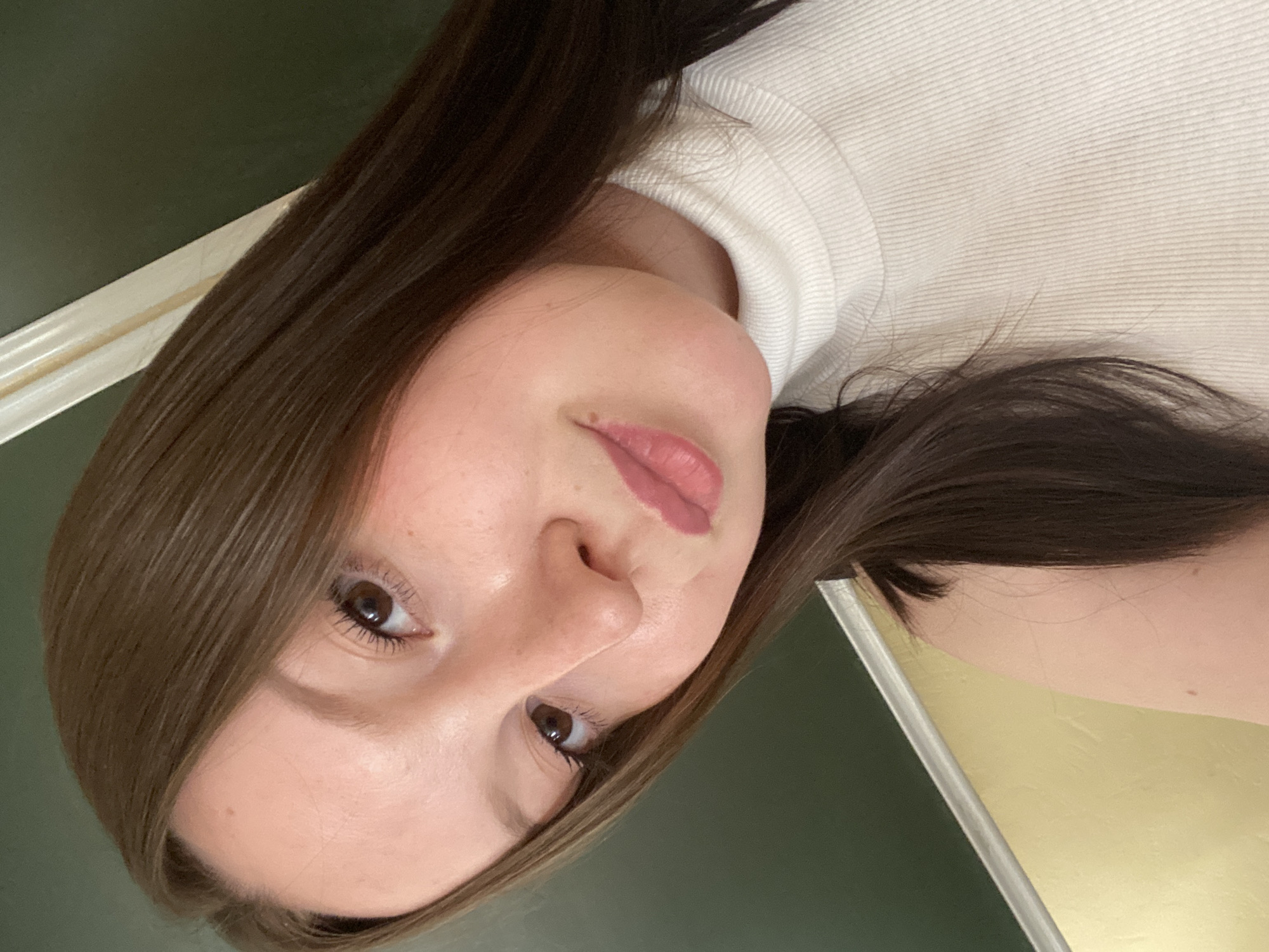 A young woman is posing for a selfie-style photograph. She has long, dark brown hair and is wearing a white turtleneck sweater. The background is a mix of dark green and light beige. It is not possible to confirm the location as Chaikovsky, Russia based on the image.