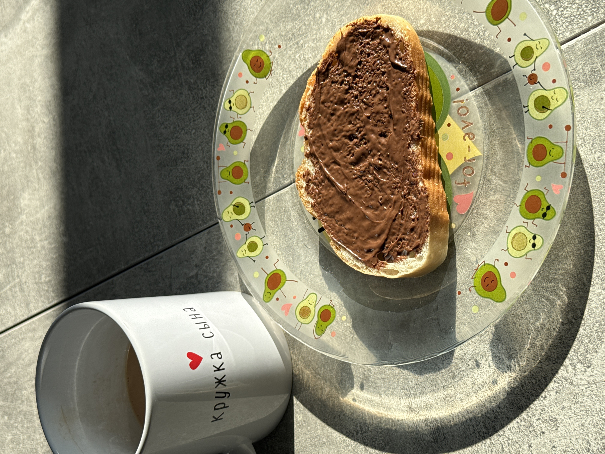 A close-up, high-angle, sunlit shot shows a white mug and a transparent glass plate with a slice of bread topped with chocolate spread. The mug, placed on the left, has a red heart and Russian text that reads "кружка сына" (mug of son). The plate, positioned to the right and slightly below the mug, is adorned with cartoon avocados and the text "for-avo". The bread slice is centered on the plate. The scene is set against a grey tiled surface, with dappled sunlight casting shadows. The image appears to be taken indoors, possibly on a balcony or near a window. There are no people or animals present in the scene. The location is identified as Lyubertsy, Russia.