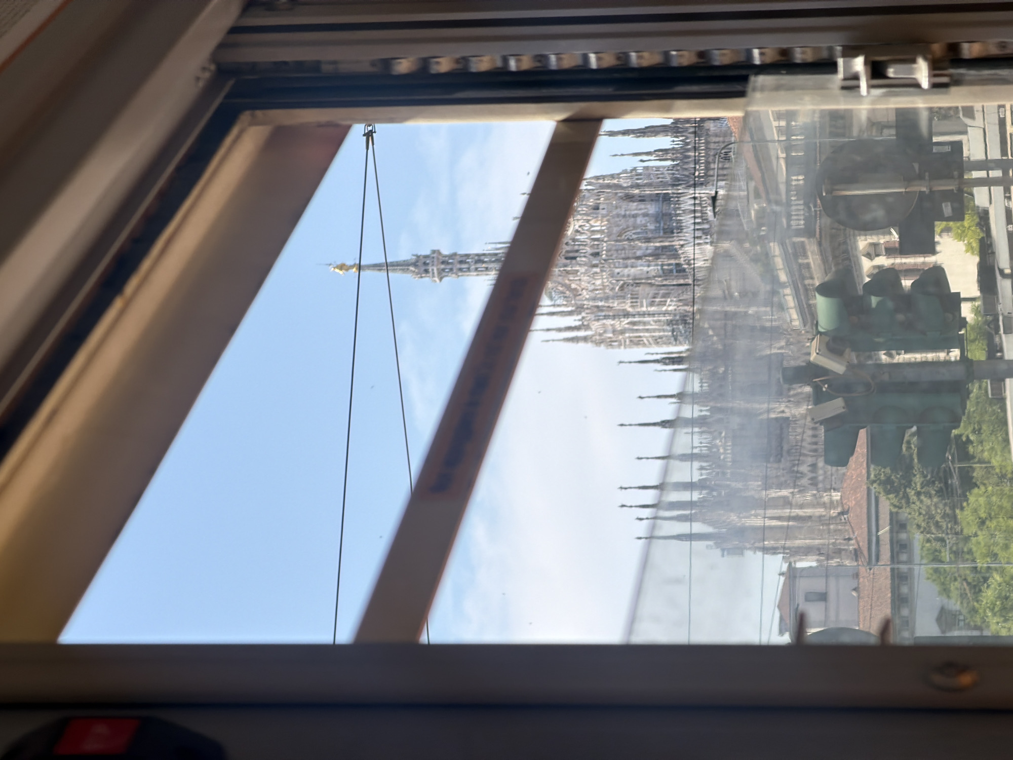 The image captures a view of the Milan Cathedral, Italy, through a window. The cathedral's spires are visible against a blue sky with scattered clouds. A traffic light and other city buildings are seen in the reflection on the window. The window frame is a light tan color and partly obscures the view. A thin wire runs vertically in front of the cathedral.