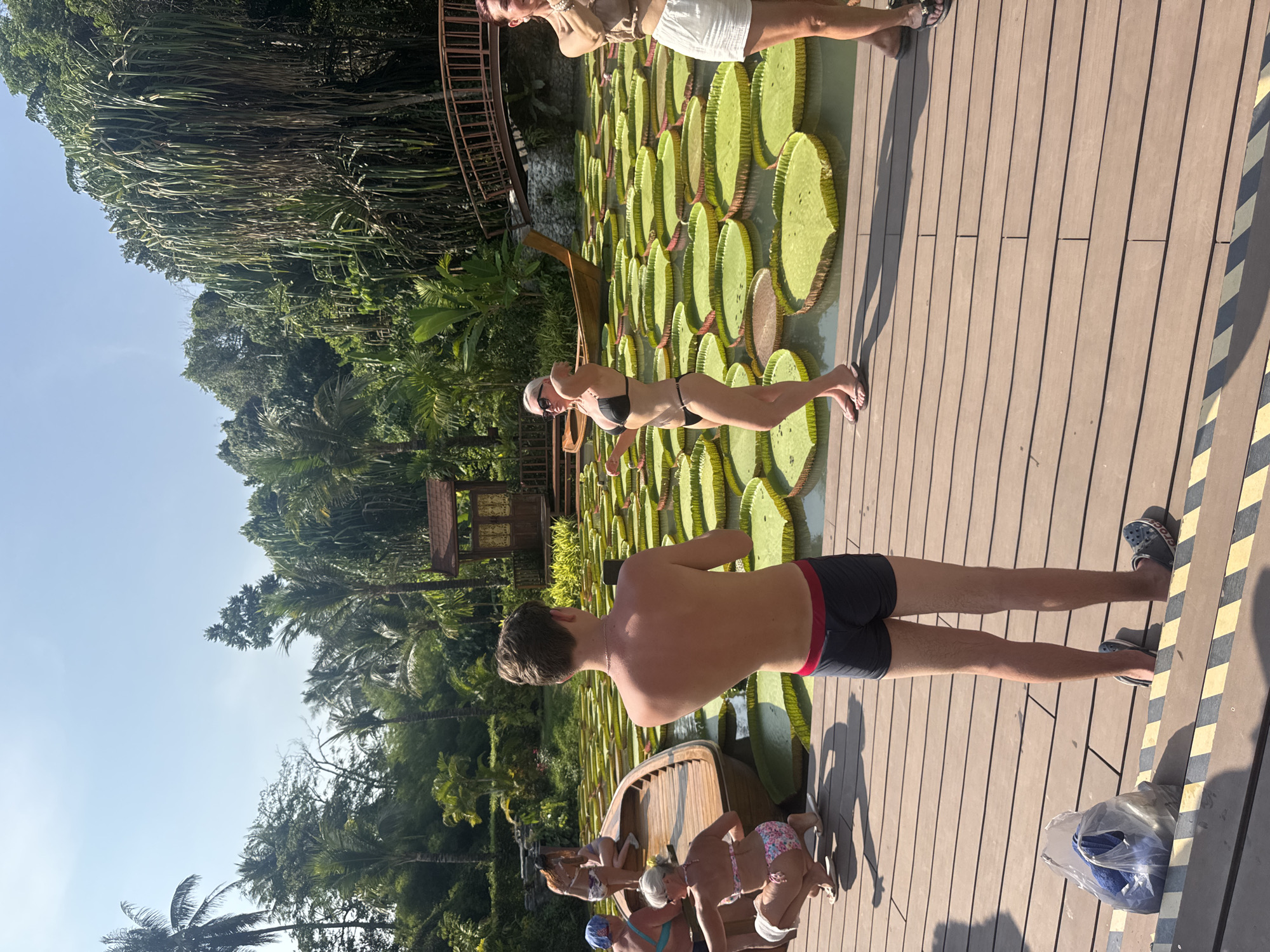 The image shows a group of people at a tourist attraction. The location appears to be a wooden deck area situated near a pond or waterway filled with giant water lilies. People are standing on the lilypads. There are also some people standing on the deck area. Trees and greenery can be seen in the background. The city is Ban Khlong Khian, Thailand.