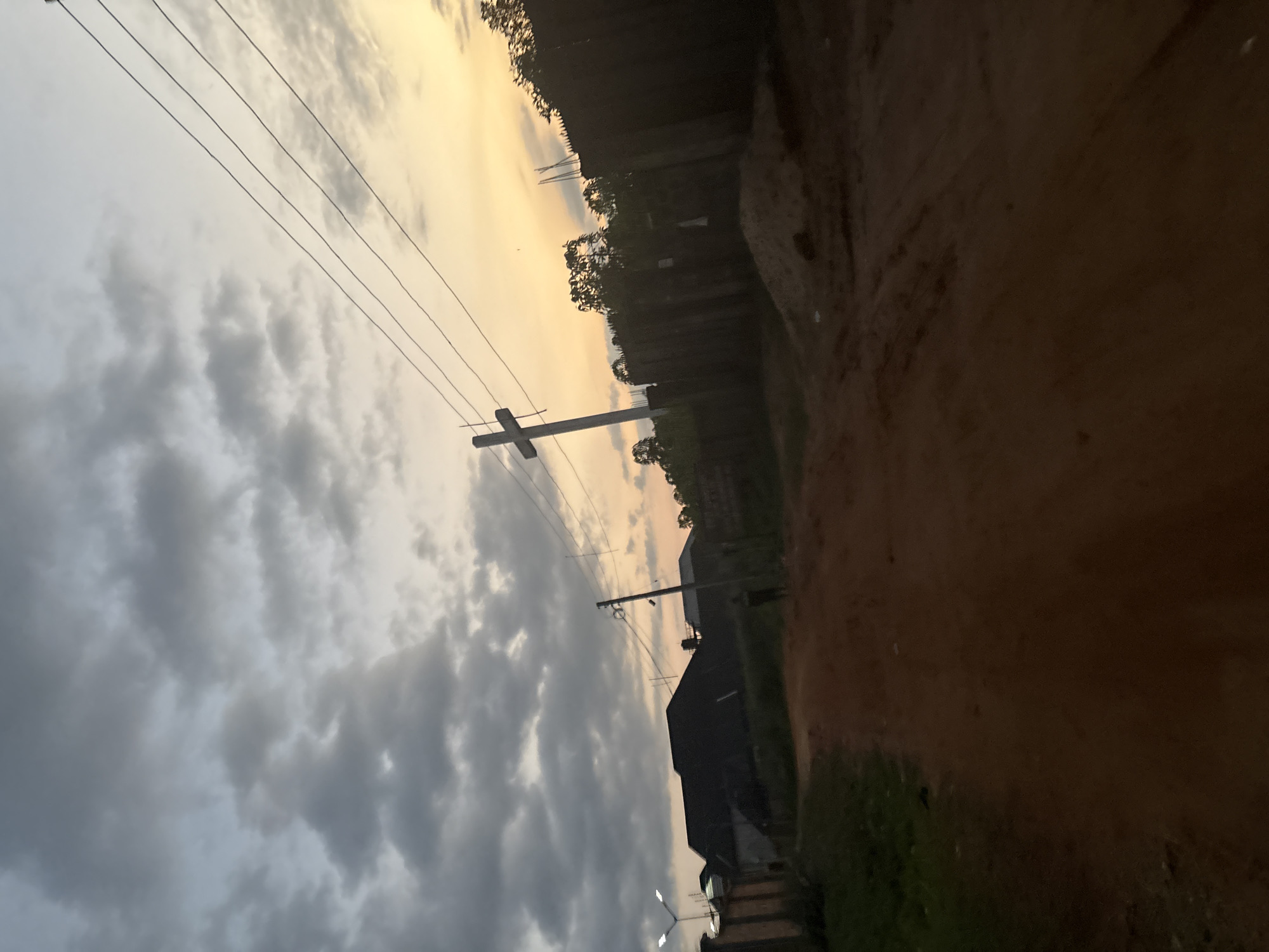 The image depicts a dirt road in the city of Obosi, Nigeria. The sky above is filled with dramatic clouds, tinged with the warm hues of a sunset. In the foreground, the reddish-brown dirt road curves towards the right, with patches of green grass visible on the left. The middle ground features a dark-roofed building, a tall utility pole with power lines, and a prominent cross on a pole. Further back, a dark fence stretches across the scene, and trees are visible behind it. A lone figure can be seen standing on the dirt road in the middle distance. The overall impression is one of a rural or semi-rural setting at dusk, with a mix of natural elements and human infrastructure.