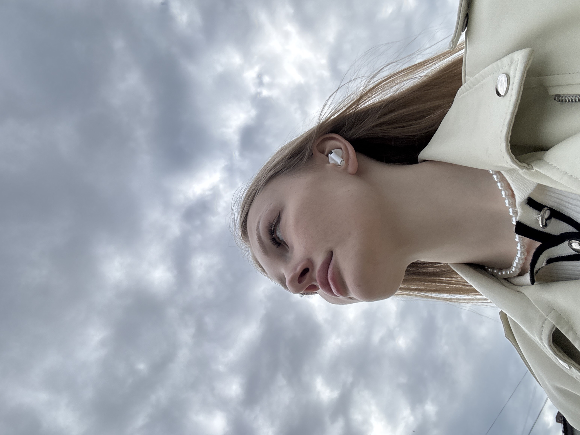 An outdoor, low-angle portrait in Perm, Russia, features a young female with long, light-colored hair, looking upwards and to the viewer's right. She is wearing a cream-colored, leather-style jacket over a white ribbed top with black and white striped detailing, accessorized with a pearl necklace. A white wireless earbud is visible in her right ear. The background consists entirely of a densely overcast sky filled with gray clouds, providing diffuse illumination. Indistinct dark shapes, possibly urban structures or utility lines, are minimally visible at the bottom-left edge of the frame.