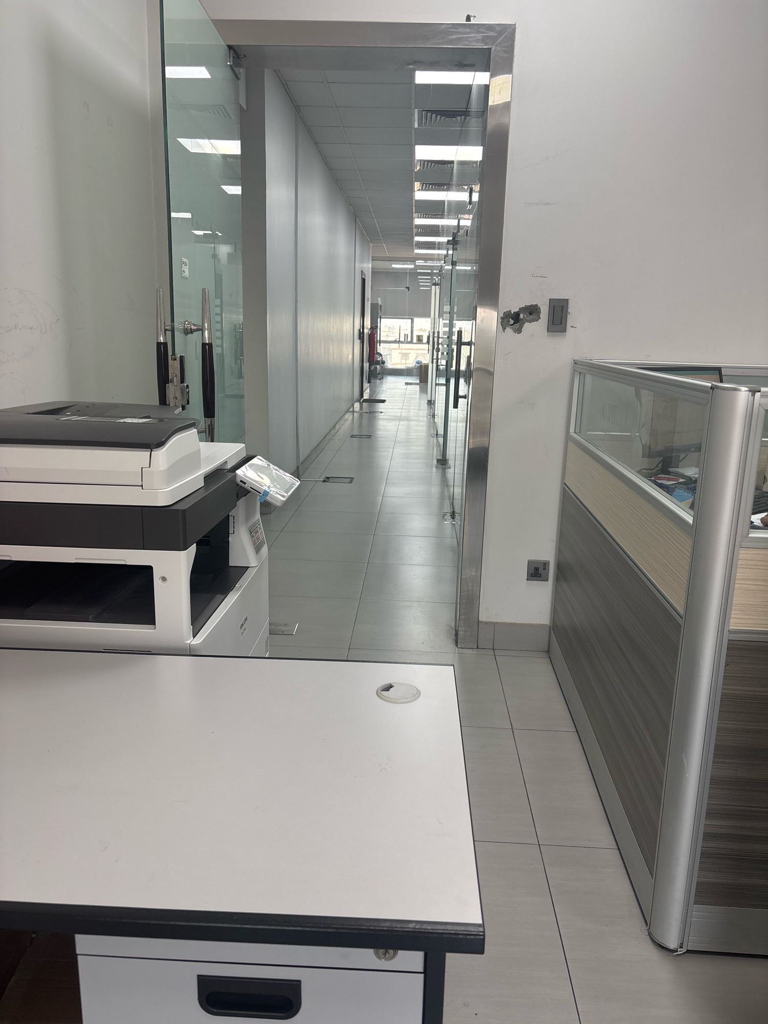The image depicts an office hallway in Dhahran, Saudi Arabia, featuring a modern aesthetic with glass partitions and white walls. In the foreground, a white desk with a black trim is visible, with a portion of a filing cabinet drawer underneath. To the left, a black and white office printer is situated on the desk. The hallway stretches into the distance, illuminated by overhead fluorescent lighting. Glass doors and cubicle partitions line the hallway, revealing glimpses of desks and office equipment within. The floor is tiled with light-colored square tiles. A gray electrical outlet is mounted on the wall to the right, near the floor.