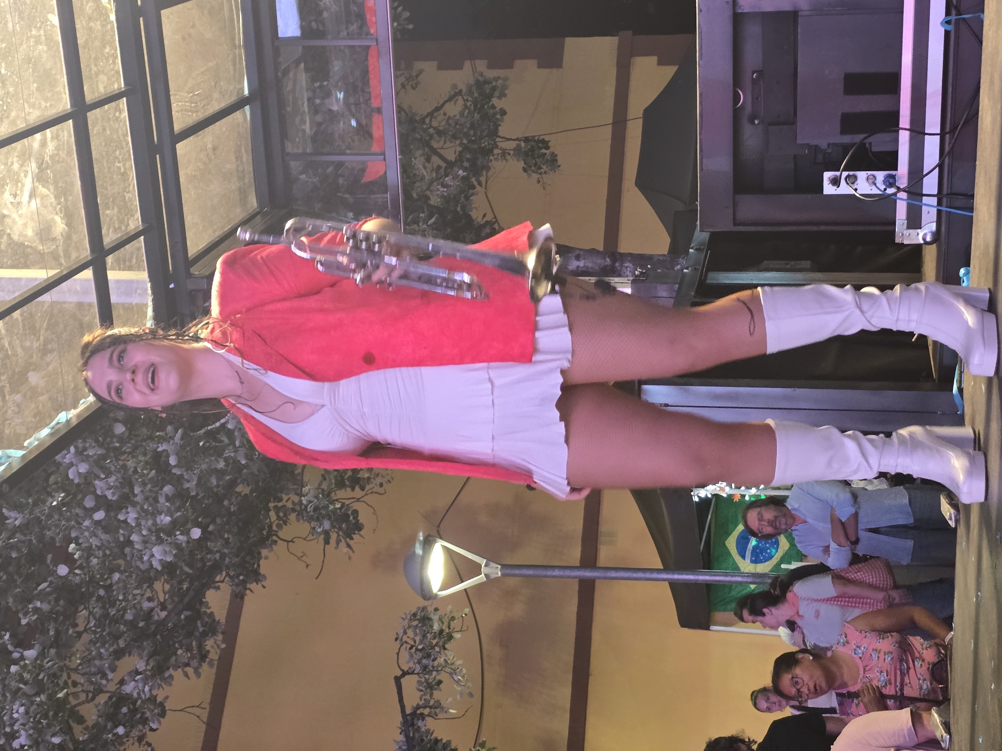 The image depicts an outdoor performance in Cornellà de Llobregat, Spain. A woman, estimated to be in her 20s or 30s, stands center-frame on a stage, holding a silver trumpet in her left hand. She is smiling and looking upwards and to her right. She wears a bright red blazer over a white, sleeveless, V-neck mini-dress with a ruffled hem, fishnet stockings, and knee-high white platform boots. Her dark hair is styled in braids or dreadlocks, and a dark, curvilinear tattoo is visible on her left lower leg, just above her boot.
To the left of the performer, a tall street light with a glowing fixture is visible. Behind the stage, a building with a light yellow/orange facade and dark brown trim is partially obscured by decorative green foliage, possibly artificial, draped across the wall. Above the stage, a metallic grid structure, likely part of a canopy or roof, is visible. On the right side of the stage, dark sound equipment or speakers are set up, with cables visible on the floor.
In the background, below stage level, an audience is visible. A Brazilian flag, displaying its green, yellow, blue, and white design, is prominently hung behind a group of spectators on the right. Among the audience members, a man with a beard and glasses, wearing a light blue collared shirt, stands with his arms crossed, looking towards the stage. Other individuals of varying ages are also present, observing the performance. The ground surface of the stage is dark.