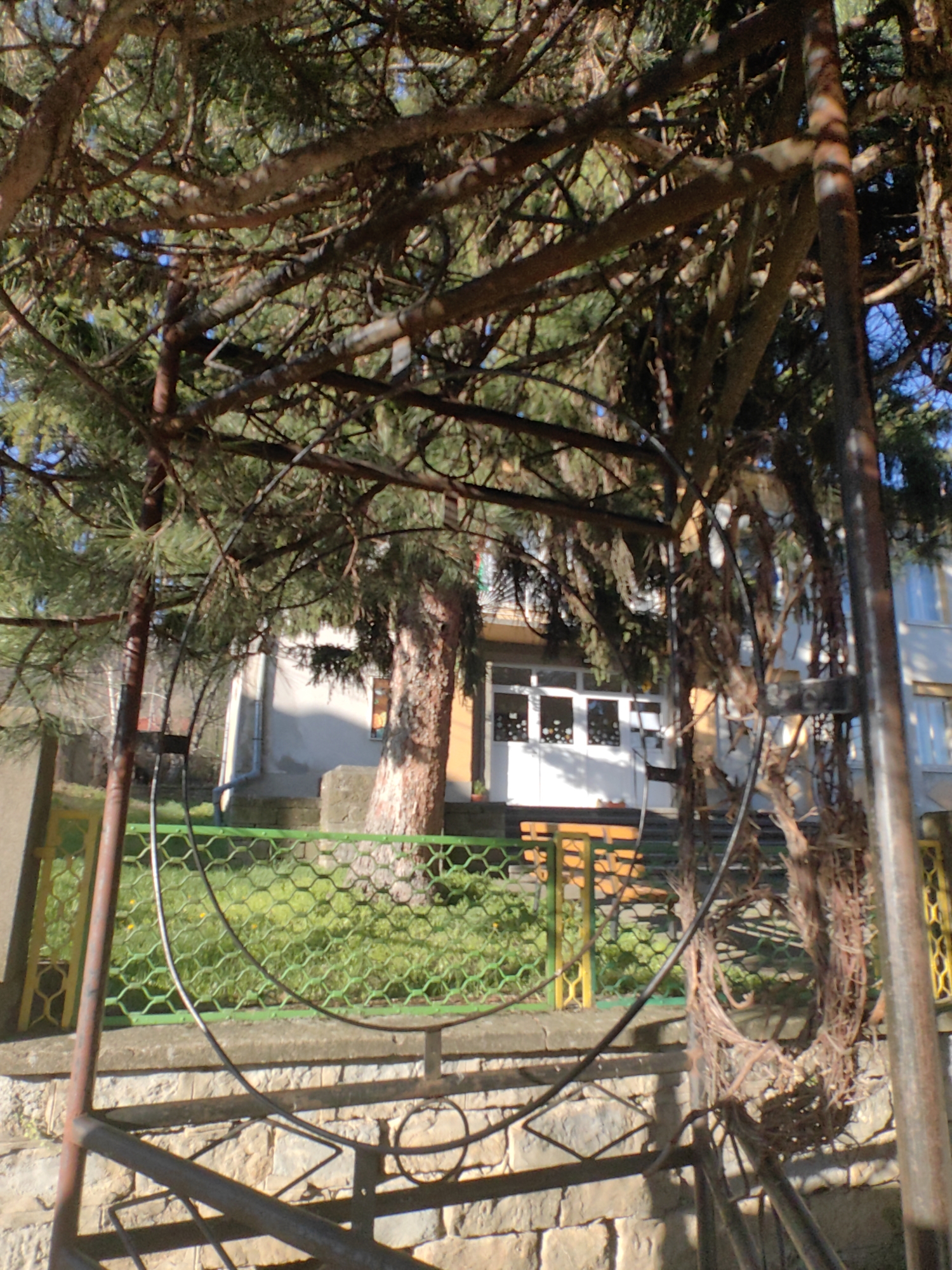 The image depicts a partially obscured view of a building behind a fence and some foliage in Hirevo, Bulgaria. A weathered, possibly rusted metal structure dominates the foreground. Beyond the metal structure and a stone wall, a green and yellow metal fence separates the foreground from a lawn. Behind the fence is a two-story building with a white facade. The building has a visible row of windows on the ground floor and an upper floor with what appears to be a balcony. There are trees both behind and partially obscuring the building, with their branches extending into the foreground. The lighting suggests a sunny day. No people are visible in the frame.