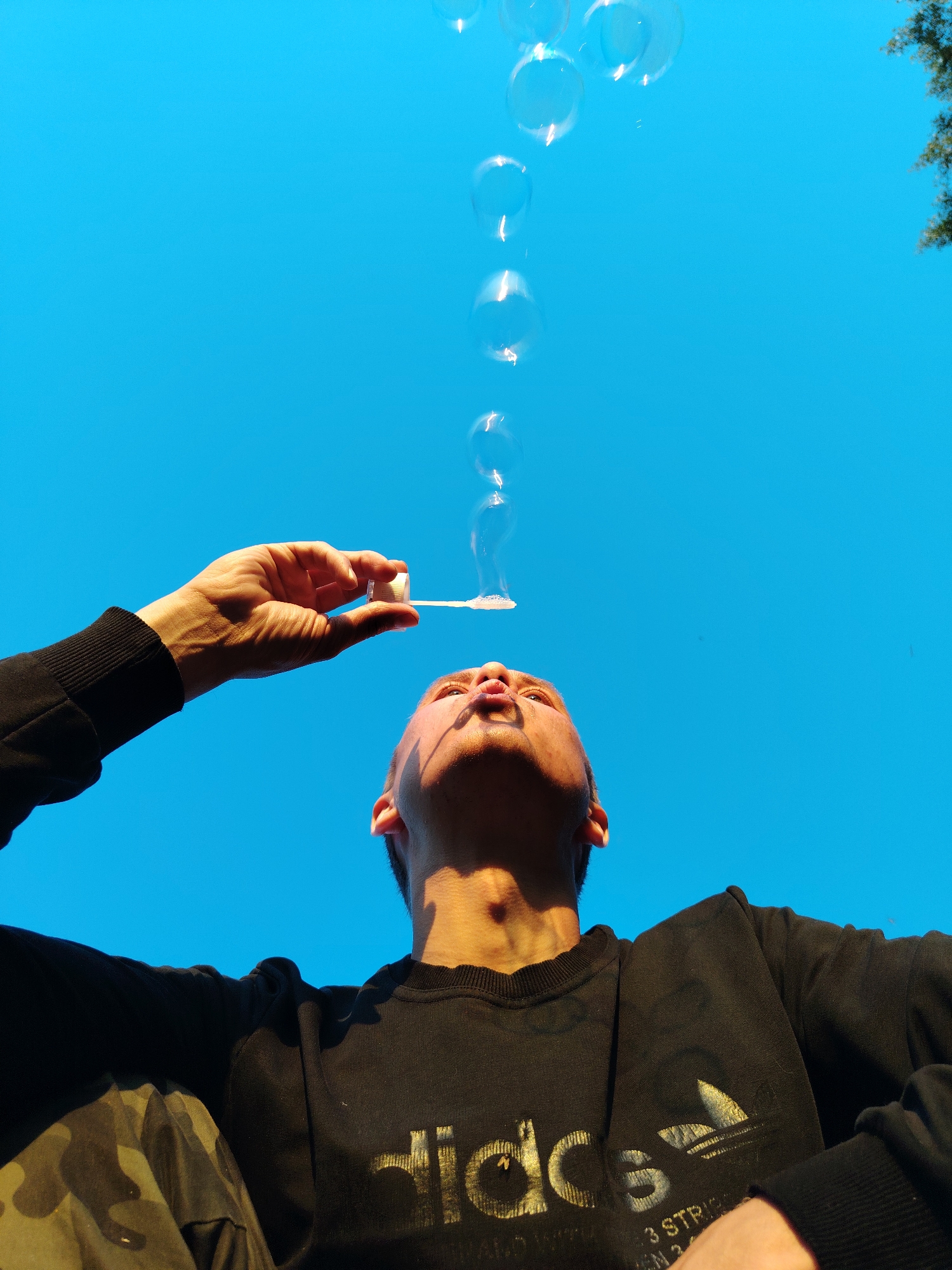 A low-angle shot captures a young person blowing bubbles against a bright blue sky. The person, seen from the chest up, is wearing a black sweatshirt with the Adidas logo visible. Their face is tilted upwards, lips pursed as they blow. A hand holds a bubble wand, with several iridescent bubbles ascending into the sky. The background is a clear, cloudless sky, with a hint of green foliage visible in the upper right corner. There is no indication of the city of Chaikovsky, Russia in this image.