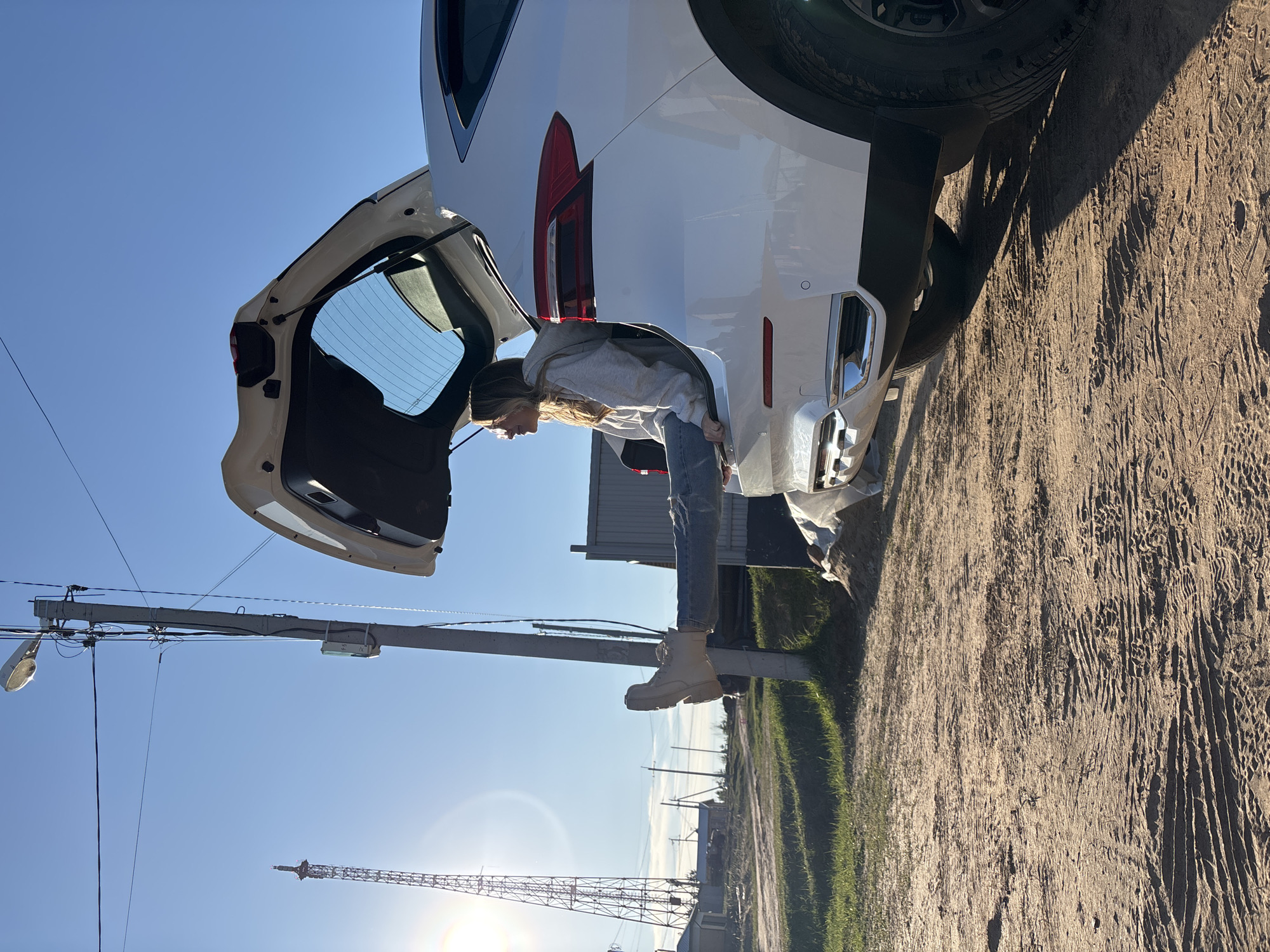 A fair-skinned woman is bent over at the open rear hatch of a white SUV parked on a dirt road on a sunny day. She has long brown hair, is wearing a light gray hoodie, blue jeans, and beige boots. A power line is visible in the left foreground against a bright blue sky. A tall radio tower is visible in the distant left. The SUV is positioned to the right. The location is Chaikovsky, Russia, based on the provided data.