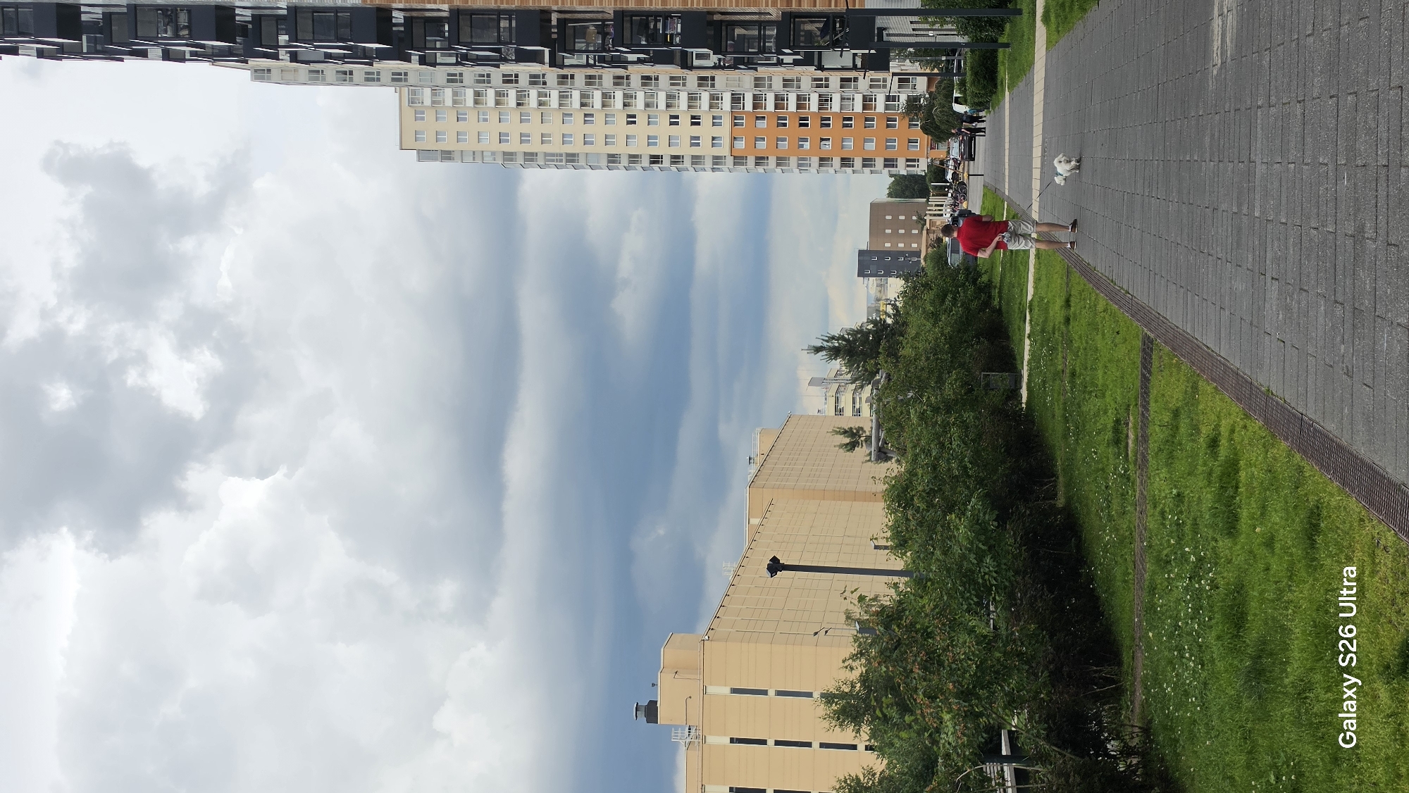 A vertical, eye-level shot captures a person walking a small white dog on a paved walkway in front of apartment buildings. The person, wearing a red t-shirt and light-colored shorts, is seen from behind, holding a leash attached to the dog, which is walking ahead. The walkway is constructed of gray paving stones and bordered by a patch of green grass with small white flowers. To the left, a dense row of green bushes separates the walkway from more grassy areas and a large, beige apartment building with dark windows. To the right, a very tall, modern apartment building with numerous balconies and balconies features a facade of cream, orange, and dark gray panels. In the background, other residential buildings and trees are visible under a heavily overcast, gray sky. The image is tagged with "Galaxy S26 Ultra".