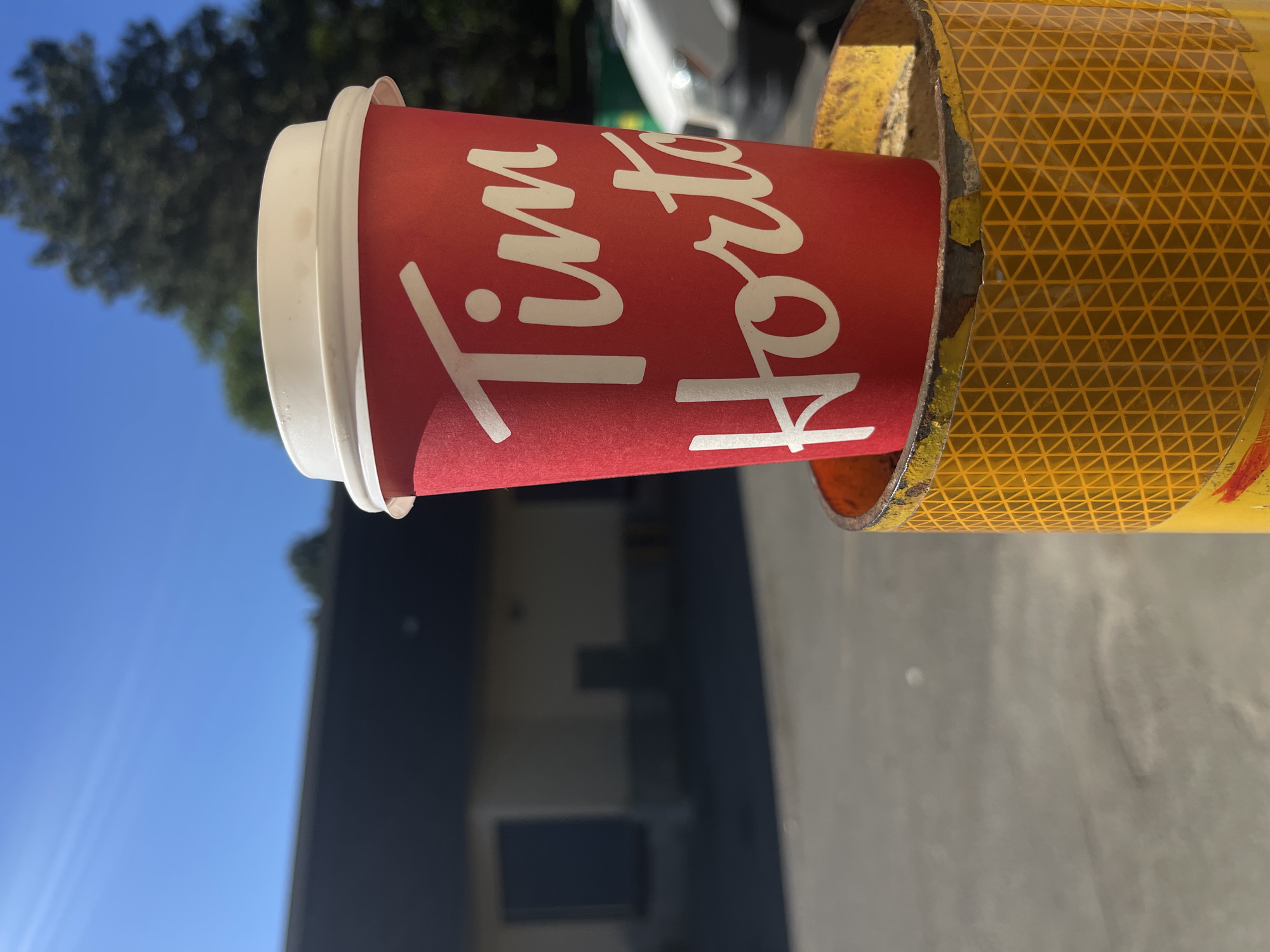 A Tim Hortons coffee cup sits inside a yellow and orange pipe. The cup is red with white lettering. The background includes a building, tree, and blue sky. The location is unknown, but the brand on the cup indicates it is likely in Canada. Given the prompt, this image was likely taken in Ottawa, Canada.