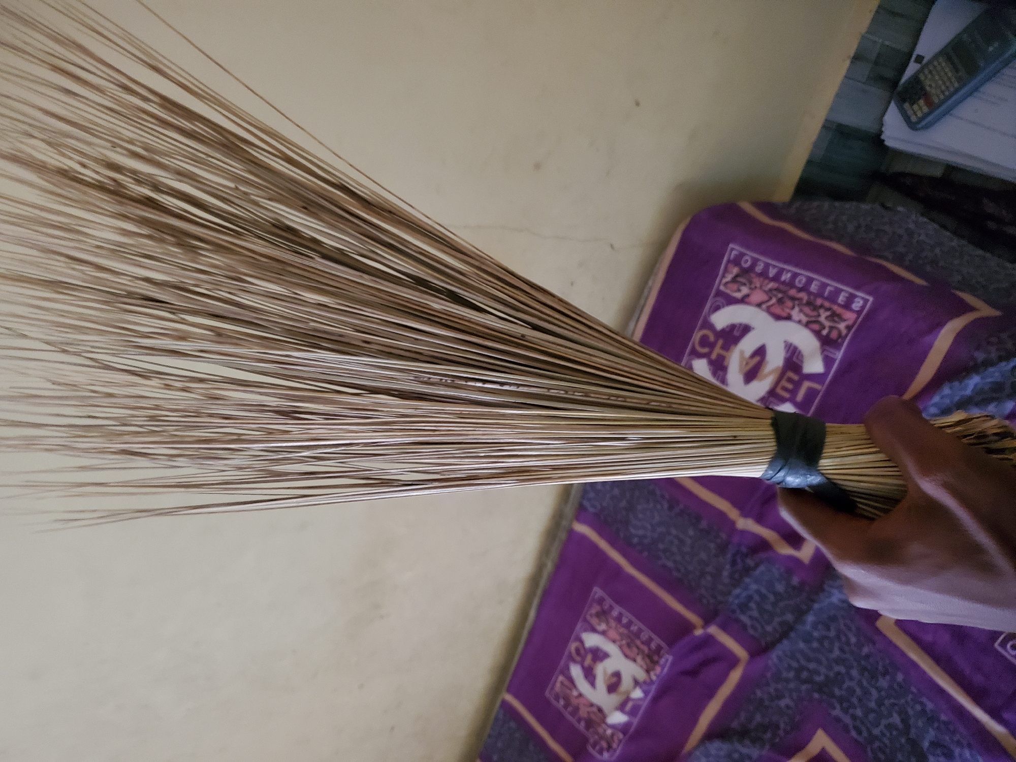 The image shows a close-up of a hand holding a broom made of dried natural fibers. The broom is tied with a dark band around its base. In the background, a bed covered with a purple textile patterned with gold and dark gray designs is visible. The wall behind the bed is a plain, light yellow color. A calculator and some papers are on the floor to the right. The scene appears to be indoors, likely in a residential setting in Jalingo, Nigeria.