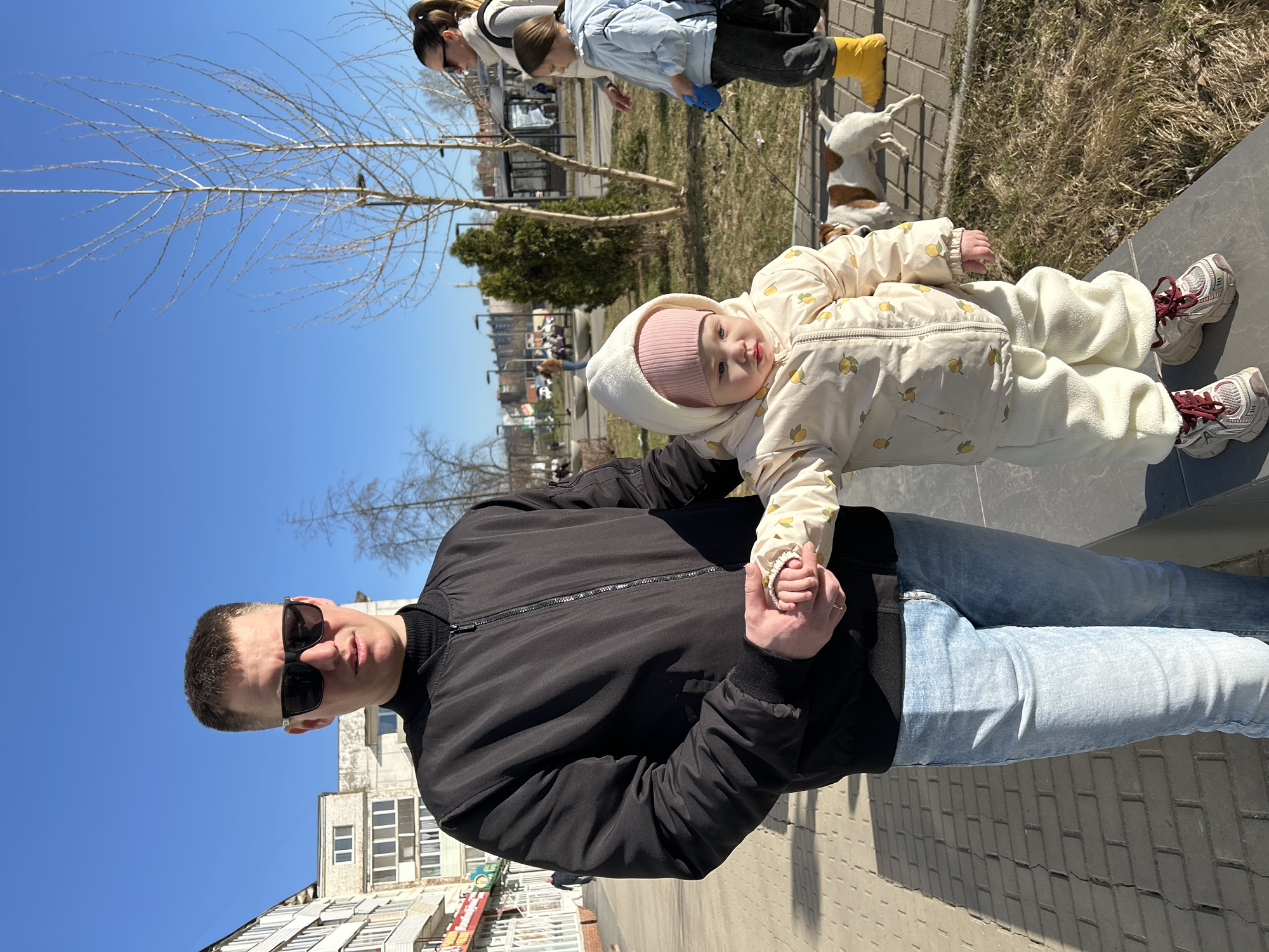 **Location:** Chaikovsky, Russia.
**Scene:** An urban outdoor scene. A man stands on a paved walkway, holding a baby. Behind them is a park area with leafless trees, grass, and other people walking a dog.
**People:**
- **Man:** Wearing sunglasses, a black jacket, and light blue jeans.
- **Baby:** Dressed in a white hooded jumpsuit with a pattern of yellow lemons.
- **Other People:** In the background, a woman, child, and dogs can be observed on the pavement.
**Objects:**
- Tree
- Pathway
- Buildings
- Park area
- Dogs
**Actions:**
- The man holds the baby's hand.
- People walk the dogs in the background.
**Interactions:**
- The man is directly interacting with the baby, holding its hand.
**Environment:**
- Clear, sunny day with a blue sky.
- Urban landscape with buildings in the background.