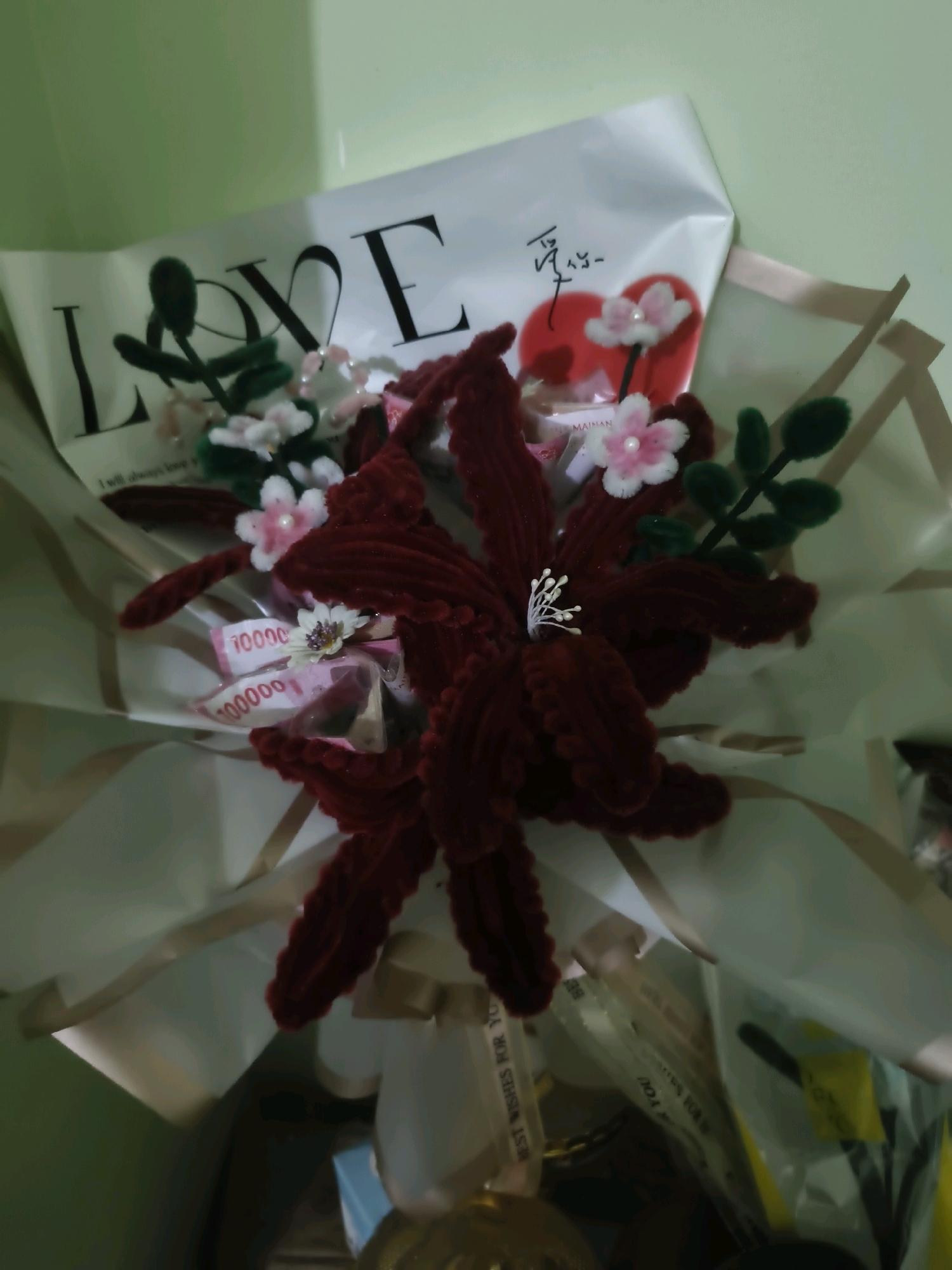 A bouquet of artificial flowers, primarily in deep red and pink hues, is presented in a light-colored paper wrap. The bouquet features a prominent dark red, five-petaled flower as the centerpiece, with smaller pink and white flowers scattered throughout. Green artificial leaves are also interspersed among the flowers. Some Indonesian currency, specifically banknotes with "10000" visible, is incorporated into the arrangement, folded and tucked between the flowers. The wrapping paper has "LOVE" printed in large black letters on the left side, and a red heart with Chinese characters and smaller text like "MAINAN" on the right. The bouquet is partially obscured by layers of light-colored, translucent paper and ribbon. The background is a light green wall. The image appears to have been taken indoors.