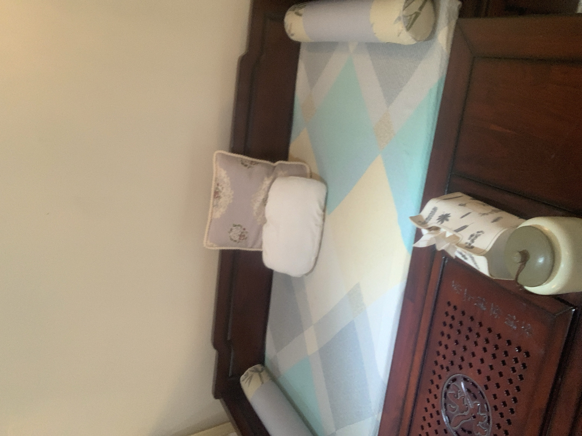 A close-up, head-on view captures the top of a dark wooden daybed, adorned with a patterned mattress in shades of white, blue, and yellow. Two cylindrical pillows, one on the left and one on the right, are placed on the edges of the bed. In the center, a small white pillow sits in front of a larger, pale purple decorative pillow with a floral pattern. To the right of the decorative pillow, a white cylindrical object with a green lid sits on the wooden frame of the daybed. The background is a plain, light-colored wall. The scene is indoors and appears to be a bedroom or living area. No specific location in Abuja, Nigeria is identifiable from the image.