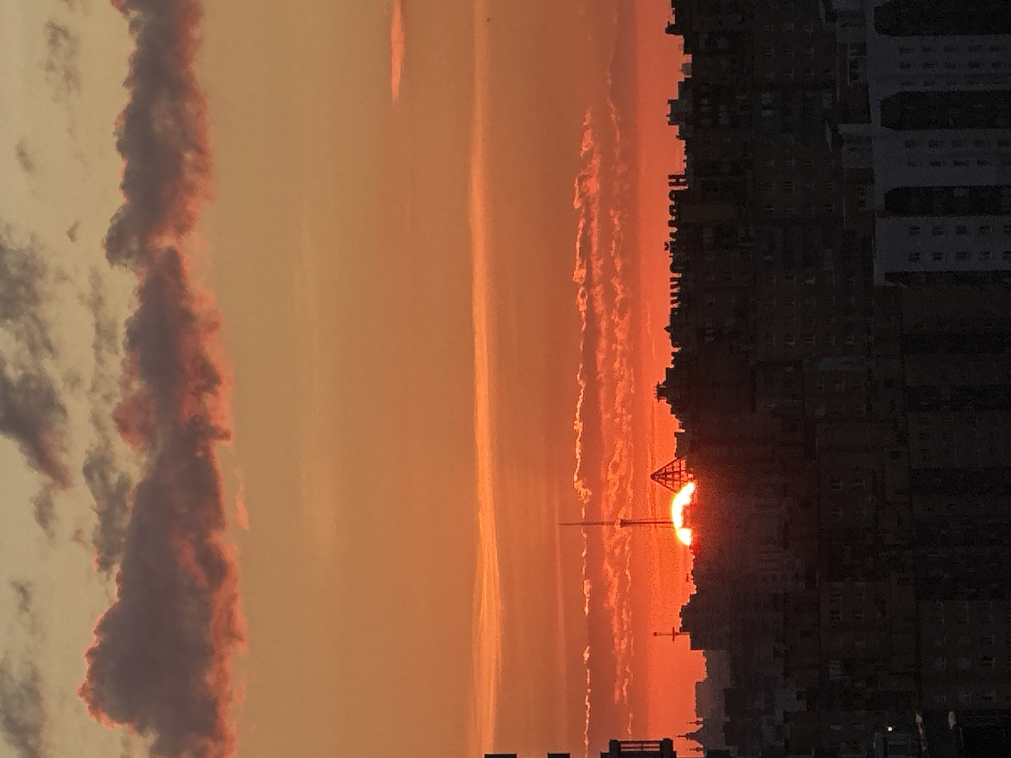 The image captures a sunset over the city of Dnipro, Russia. The sky is a vibrant gradient of orange and red, with streaks of pink and purple clouds. The sun, a bright orange orb, is partially obscured by the horizon, casting a warm glow over the cityscape. Silhouetted against the colorful sky are the rooftops of numerous buildings, appearing as dark shapes. A prominent tall antenna structure rises above the buildings, with a distinctive pyramid-shaped roof visible on another structure. The lower portion of the image is dominated by the dark silhouettes of apartment buildings, their windows reflecting the fading light. A few faint signs with Cyrillic lettering are visible on the top of some buildings. The overall impression is a dramatic and atmospheric urban sunset.