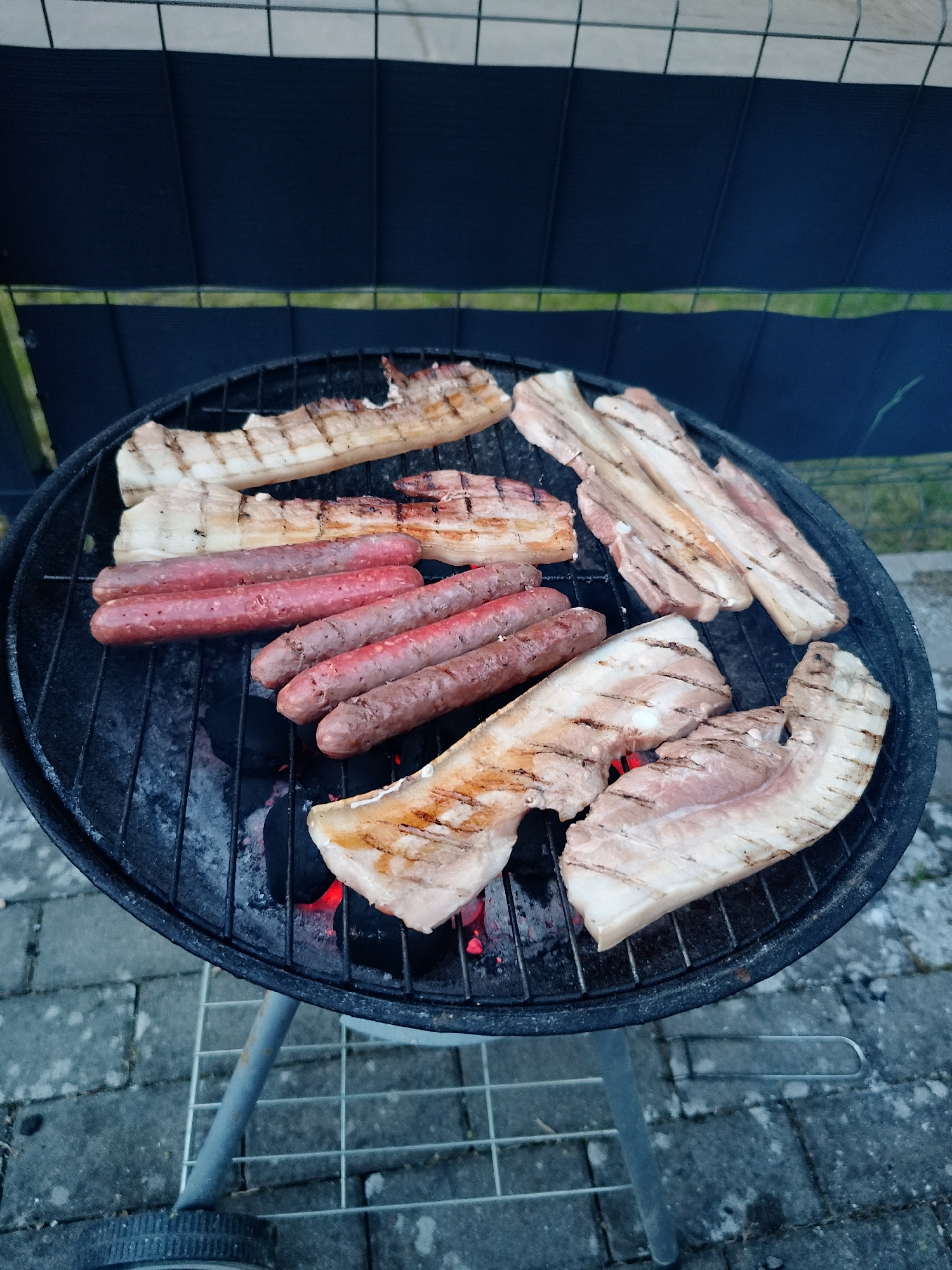 A charcoal grill is loaded with sausages and what appears to be bacon or pork belly. The grill is black, and the coals are glowing red underneath the meat. The grill is sitting on a stand with metal legs. The ground below is paved with gray bricks. In the background, there is a black fence with a thin, light green strip above it. The location appears to be outdoors, possibly a backyard, in an unknown city.