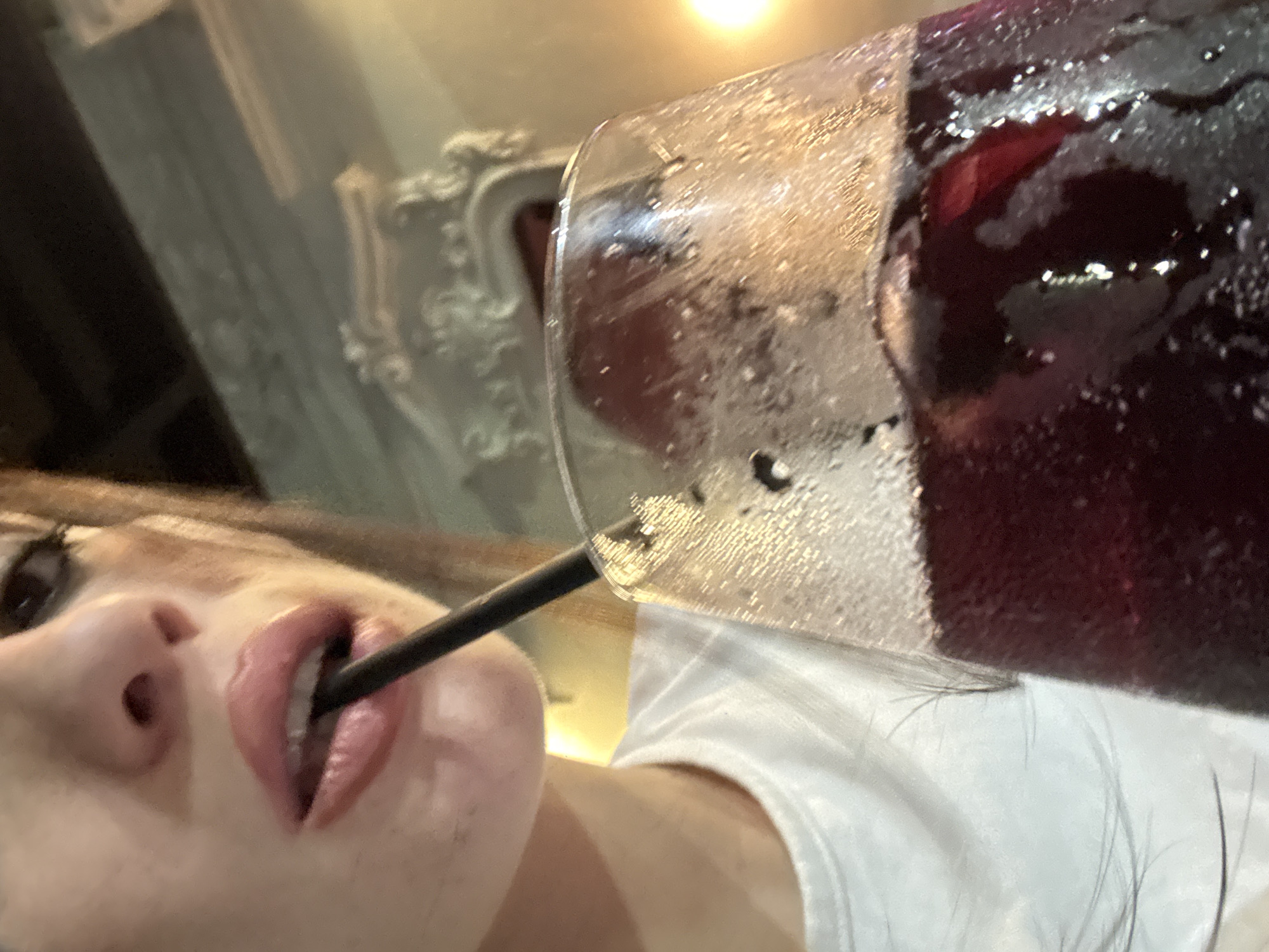 A close-up, low-angle shot features a woman drinking from a glass filled with a dark red beverage through a black straw. The glass is covered in condensation. Part of the woman's face is visible, showing her nose, mouth, and one eye. She is wearing a white shirt. The background is blurred, showing ornate architectural details. The location is not discernible from the image alone.