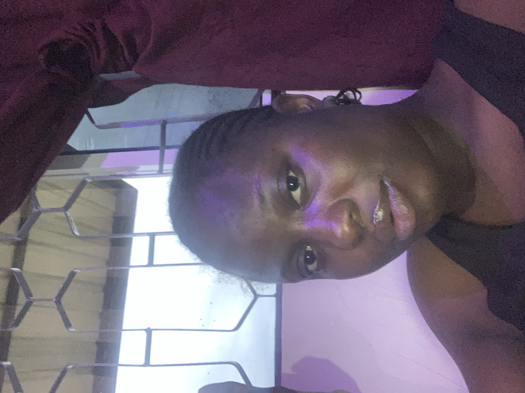 A close-up, self-portrait photograph shows a dark-skinned woman with braided hair wearing a black top. She is positioned in front of a window with a security grate. The background includes a blurred ceiling and a dark red curtain. The lighting in the scene is a mix of natural light from the window and purple ambient lighting, which casts a glow on her face and the wall. The woman's face is tilted slightly towards the camera, and her lips are parted. The overall impression is that of a casual, indoor portrait.