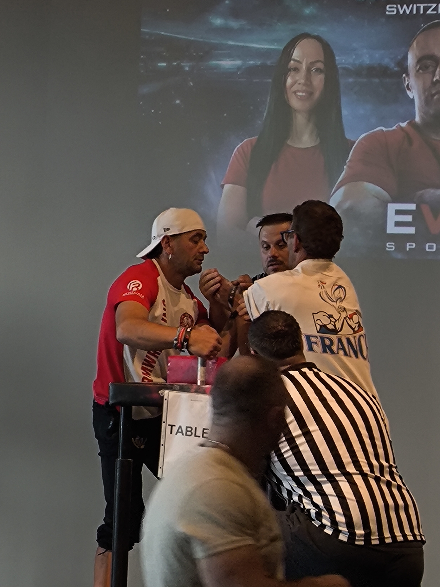 The image shows a group of people, likely at an arm wrestling event. In the foreground, two men are engaged in an arm wrestling match, their hands clasped together over a table. One man wears a red and white shirt with a white baseball cap, and the other wears a white t-shirt with a graphic design on the back featuring a French flag and arm wrestling imagery. A person in a black and white striped shirt, possibly a referee, stands behind them. In the background, a large screen displays images of a woman and a man, with text that includes "SWITZERLAND" and "EV SPON". The event appears to be held indoors with a plain gray wall as the backdrop. The presence of the text "SWITZERLAND" on the screen strongly suggests the location is in Switzerland. The setting resembles a sports competition or tournament.