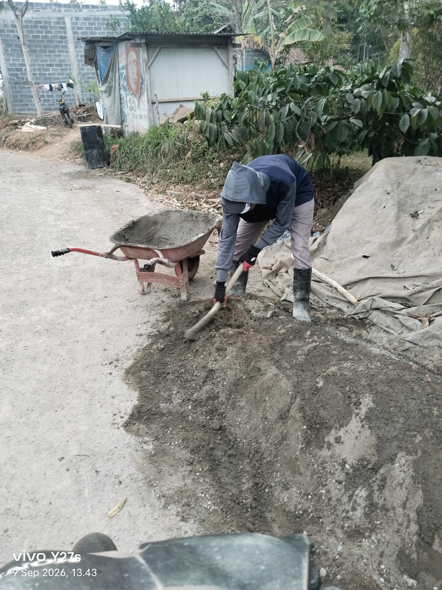 A person is using a shovel to mix cement and aggregate on the ground. The person is wearing a hooded jacket, light-colored pants, black boots, and gloves. A red wheelbarrow filled with cement is next to them. The scene is outdoors, with a dirt and gravel ground. In the background, there are some buildings, trees, and a large tarp covering a mound of material. The photo appears to have been taken in a rural or semi-rural area. The timestamp at the bottom of the image indicates it was taken on September 9, 2026, at 13:43.