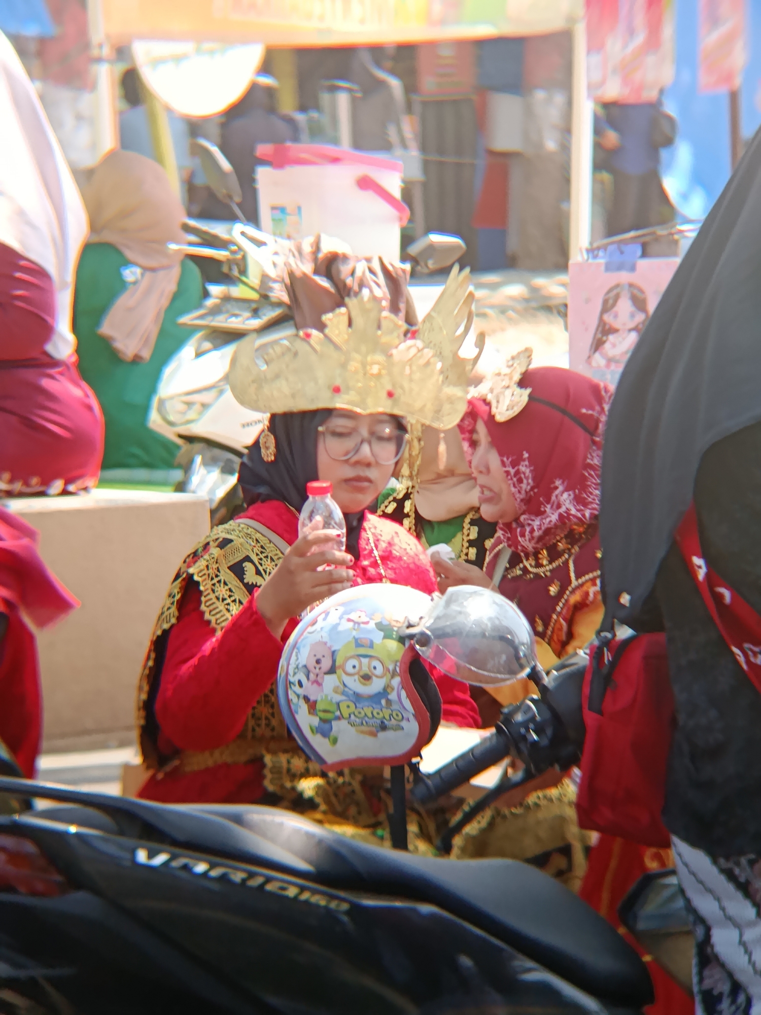 In the city of Desa Kebon Manis, Kecamatan Cilacap Utara, Indonesia, two individuals are depicted in the foreground, positioned outdoors in what appears to be a public area. The individual on the left, wearing traditional attire with a gold headdress and a red garment with ornate gold embroidery, is holding a clear plastic bottle of water. She has glasses and is looking downwards. To her right, another individual, also wearing traditional clothing including a red hijab with white detailing and a gold-adorned garment, is looking to her left with an open mouth, seemingly speaking. Both individuals are near a scooter, which is partially visible in the lower portion of the image. The scooter's handlebars and a portion of the seat are visible, along with a helmet adorned with cartoon characters on the handlebars. In the background, other people and structures, possibly market stalls, are blurred, indicating a lively, outdoor environment. The lighting suggests it is daytime, with bright sunlight casting shadows.