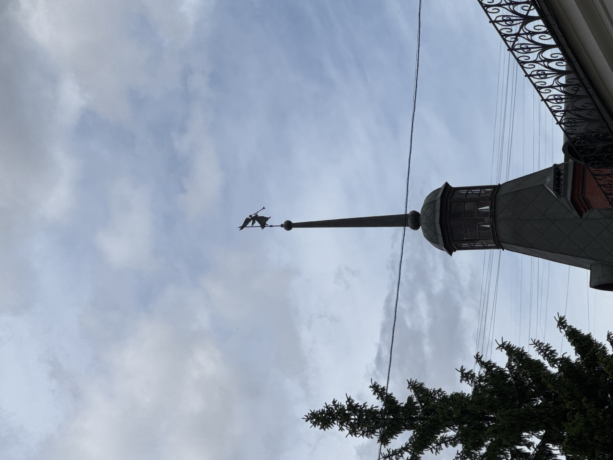 The image shows a tall, gray building with a weather vane featuring an angel playing a trumpet at the top of a spire. The building has a domed section with windows. The top right corner of the image shows ornate black wrought-iron detailing. A power line runs diagonally across the image from the top to the right side. A section of a tree with dark green foliage is visible in the bottom right corner. The sky is overcast with light gray and white clouds. The location is likely Moscow, Russia, given the architectural style of the building, which is typical of the city.