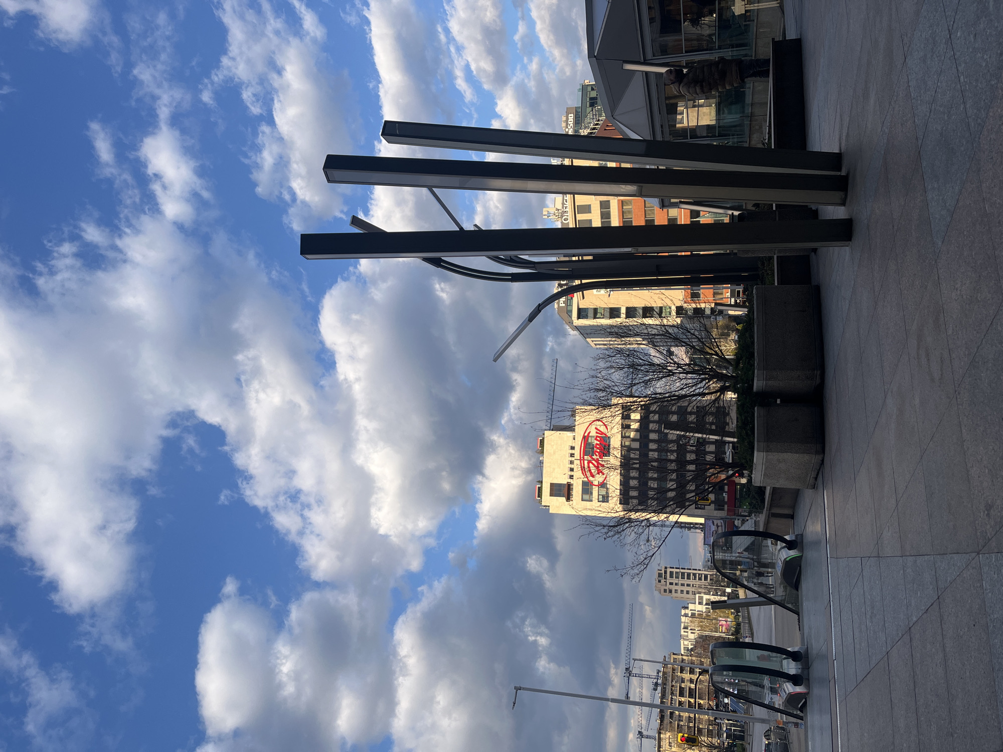 The image shows a cityscape with a blue sky and white clouds dominating the upper left portion. In the center, three dark, vertical metal beams stand prominently, part of some kind of structure. Buildings of varying heights can be seen, including one with a large, red "Fancy" sign. Bare tree branches are visible. An escalator and tiled pavement are visible in the lower right, suggesting an urban area. Construction cranes are evident in the distance. The image appears to be taken in Sofia, Bulgaria based on the "Fancy" sign which is located on a hotel in Sofia.