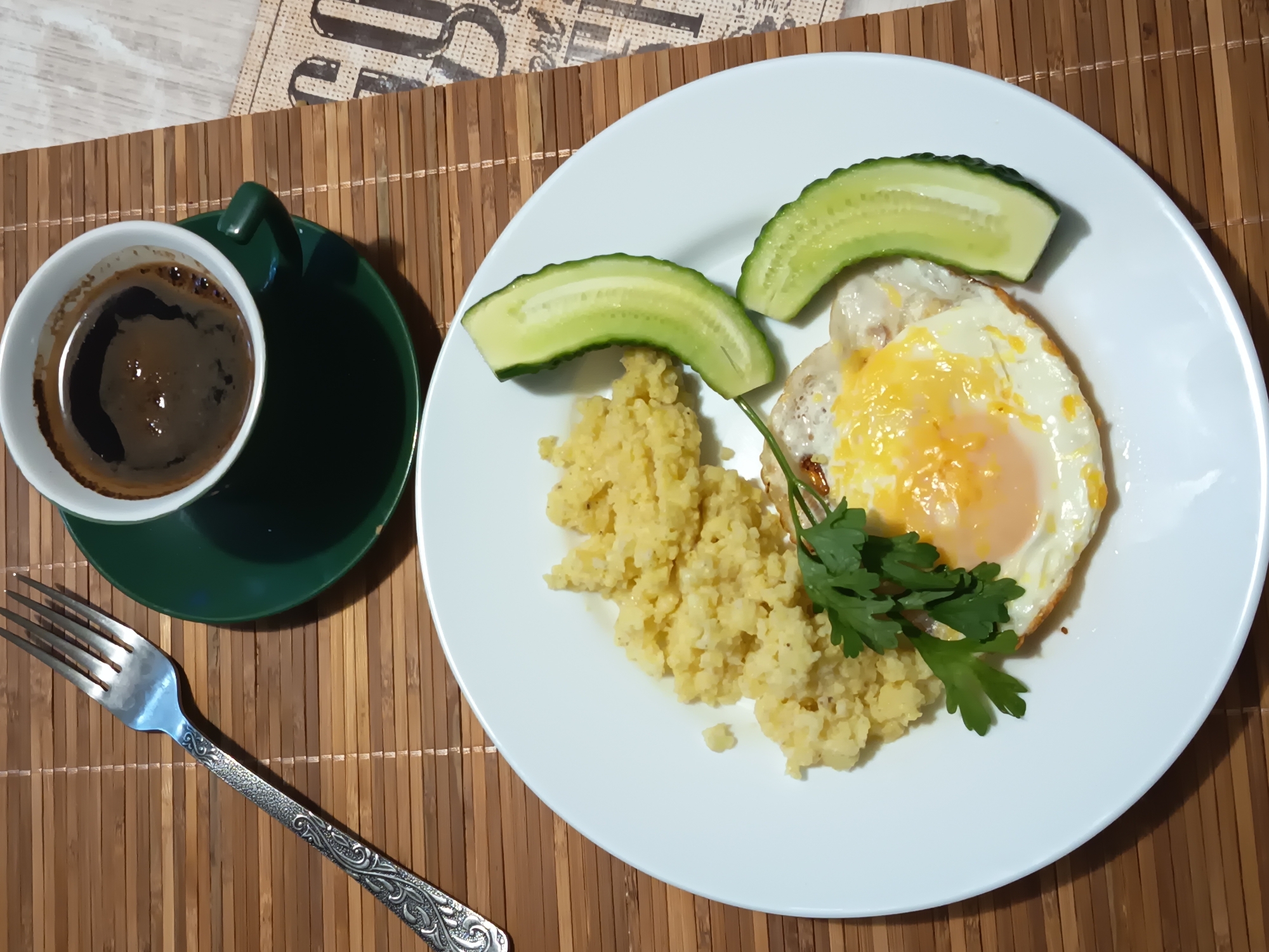 A top-down shot captures a breakfast setting on a bamboo placemat. The image features a white plate holding a fried egg, a portion of couscous, two slices of cucumber, and a sprig of parsley. To the left of the plate, a white cup filled with dark coffee sits on a dark green saucer. A silver fork with an ornate handle rests on the placemat near the cup. The background shows a hint of a newspaper. The scene appears to be indoors, likely in a home or cafe, in Izmail, Ukraine. No people are visible in the image.