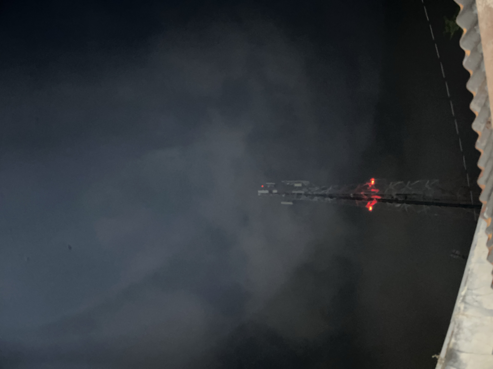An image taken at night shows a cellular tower against a dark, cloudy sky. The tower has several antennas and red warning lights visible. In the foreground, a portion of a corrugated roof or wall is visible, suggesting an elevated viewpoint. The scene appears to be located in Pekalongan, Indonesia.