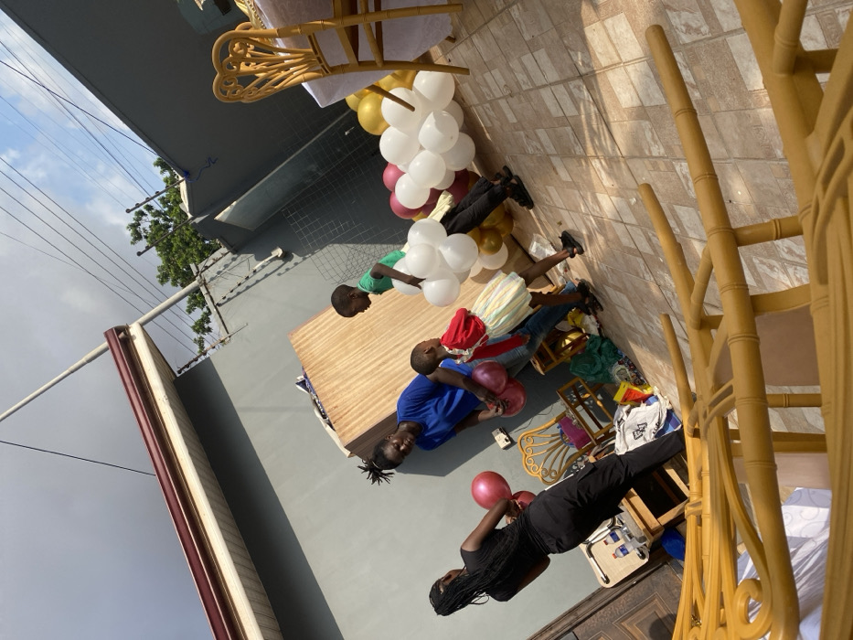 The image depicts an outdoor daytime scene at an unidentified location in Ghana. Four individuals are present: two adults and two children. On the left, a person with dark braided hair, wearing a black top and trousers, stands with their back to the viewer, holding two metallic pink balloons. Adjacent, an adult in a blue t-shirt and jeans holds additional metallic pink balloons. A child in a red top and striped skirt stands near the adult in blue. Further right, a child in a green t-shirt carries a cluster of white balloons. The ground is covered with light-colored tiles. Clusters of white, metallic pink, and gold balloons are arranged on the tiled surface, extending towards the right. Several gold-colored, bamboo-style chairs are visible, some stacked in the foreground, and one with a white-covered table on the far right. A wooden side table holds a white cooler and two plastic bottles near the left adult. A large sheet of plywood leans against a grey building wall in the background. Perimeter walls are grey, with the right wall featuring security wire along its top edge. Overhead, electrical wires are visible against a partly cloudy sky.