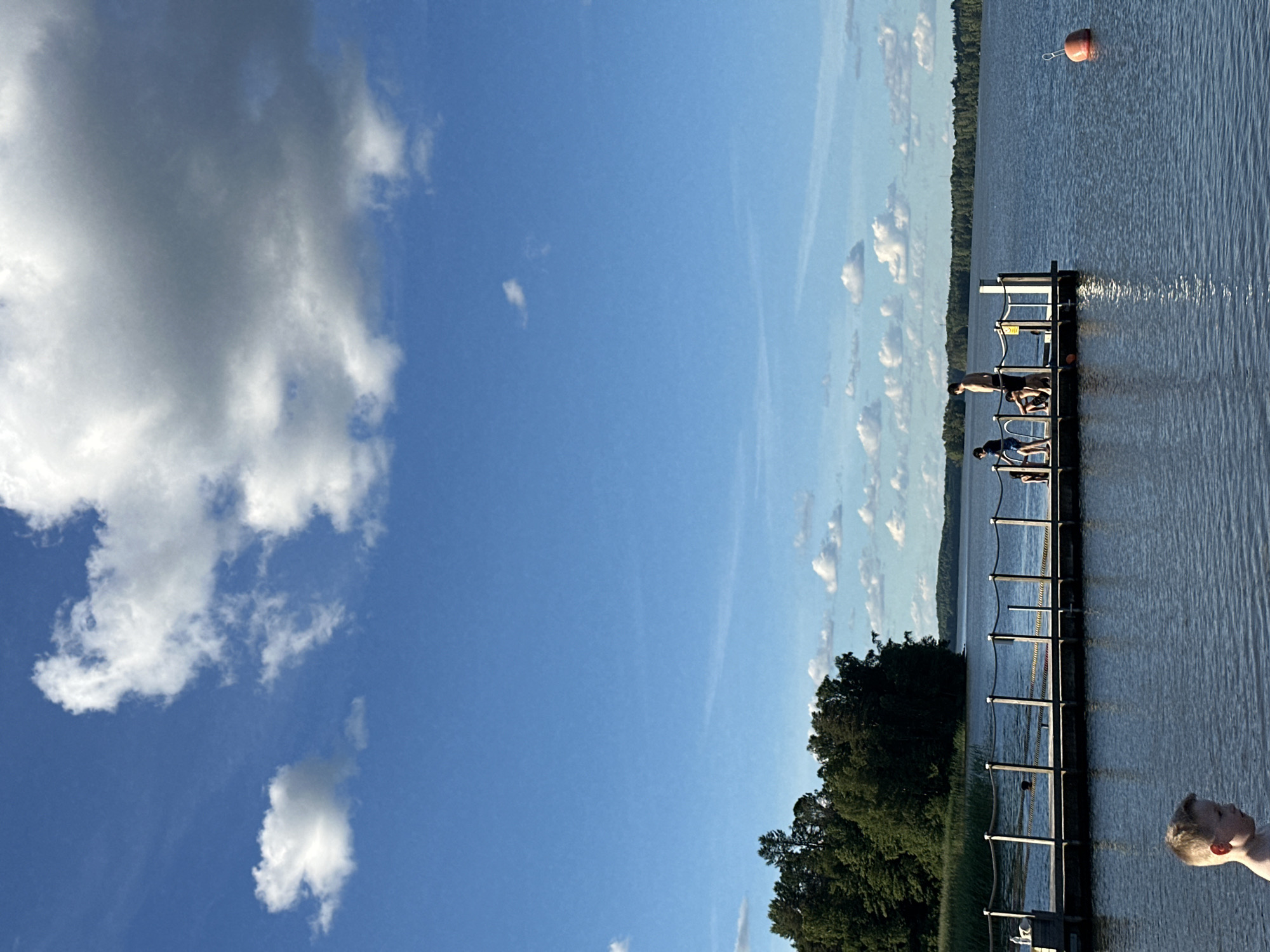 The image depicts a group of people at a lake. In the foreground, a young boy stands in the water, facing to the left. A wooden pier extends into the lake from the left side of the frame, with several people standing on it. Some appear to be children and adults. The pier is equipped with a rope railing. The lake is calm, with a few ripples on the surface. In the distance, a tree-lined shore is visible, with a clear blue sky dotted with white clouds above. A single orange buoy floats in the water to the right of the pier. The scene appears to be set in a natural outdoor environment, likely a recreational area.
