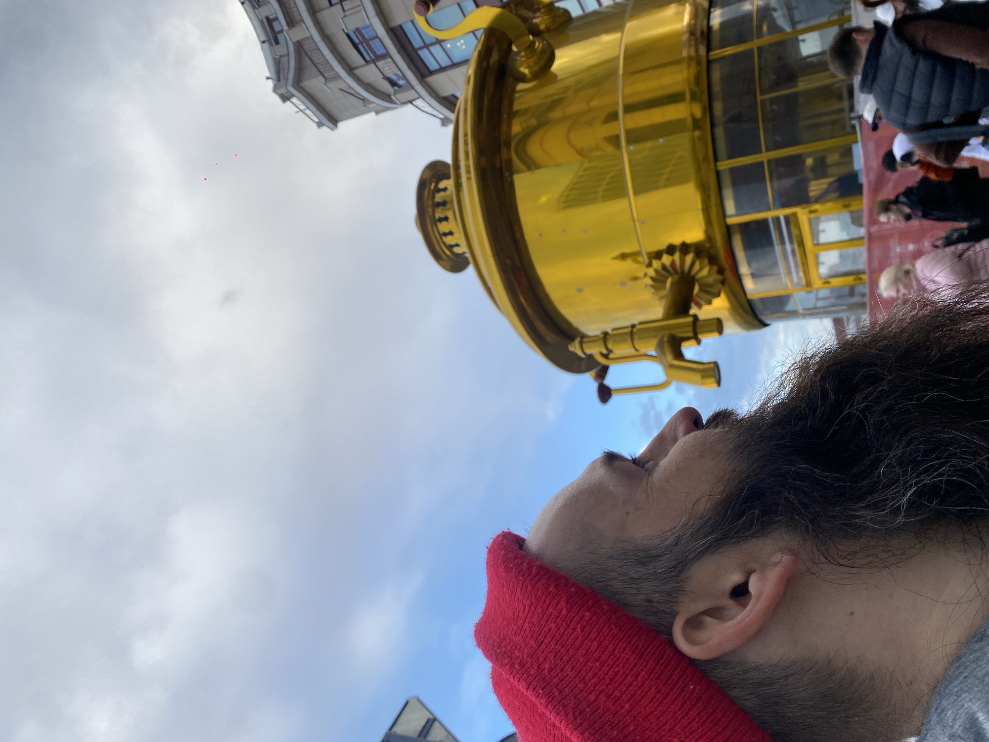 A low-angle shot depicts a man with a red beanie looking upwards towards a large, shiny golden samovar-like structure. Behind the structure, a multi-story building is visible, and several people are indistinctly seen on a platform within the samovar. The sky is partly cloudy, with patches of blue visible. The man's face is turned in profile, and his beard and ear are clearly visible. The photo seems to have been taken in Moscow, Russia, likely near the Izmailovo Kremlin where a similar structure is known to exist.
