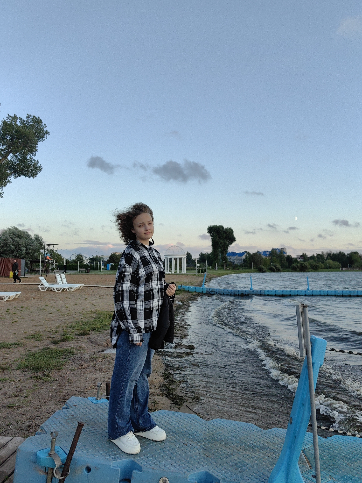 A young woman with curly brown hair is standing on a blue floating platform at the edge of a body of water. She is wearing a black and white checkered shirt, blue jeans, and white sneakers. She is holding a black jacket in her right hand. The background shows a sandy beach with white lounge chairs, some trees, a lifeguard tower, and a white gazebo. In the distance, there are buildings and a tree shaped like a heart. The sky is a pale blue with some clouds and a crescent moon. The water is rippling and there is a blue floating barrier in the water. The location is a beach in the city of Myadzyel, Belarus.