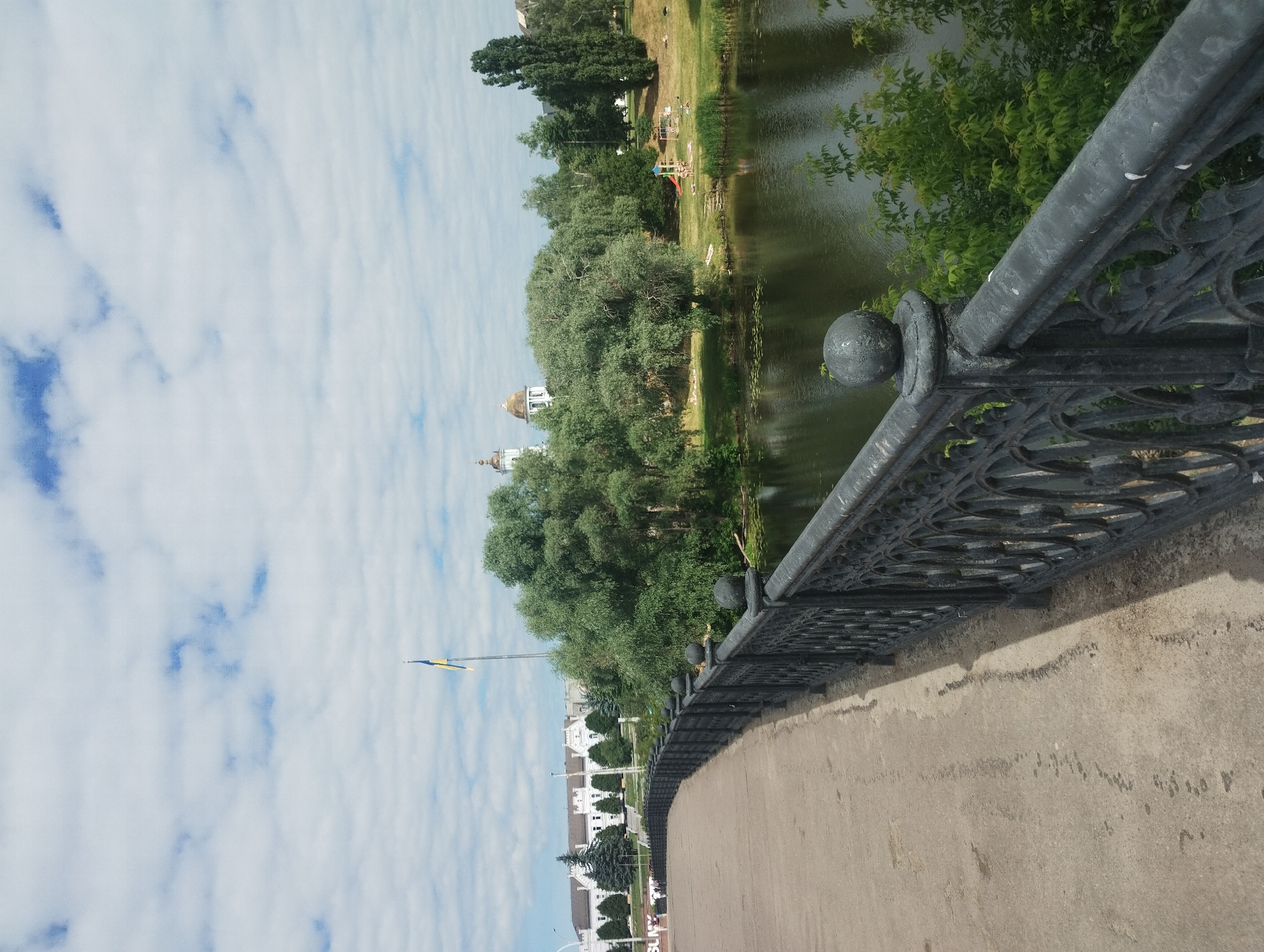 The image shows a view from a bridge overlooking a body of water, with a park area on the opposite bank and a large white building in the background. The bridge has a concrete walkway and a decorative black metal railing. In the distance, a flagpole with the Ukrainian flag is visible. The park area features lush green trees, a grassy lawn with several people sunbathing, and a children's playground with a red slide. Two church domes are also visible behind the trees. The sky is partly cloudy with bright white clouds against a blue sky. The water is dark and calm, reflecting the surrounding greenery. The overall scene suggests a warm, sunny day in a city park.