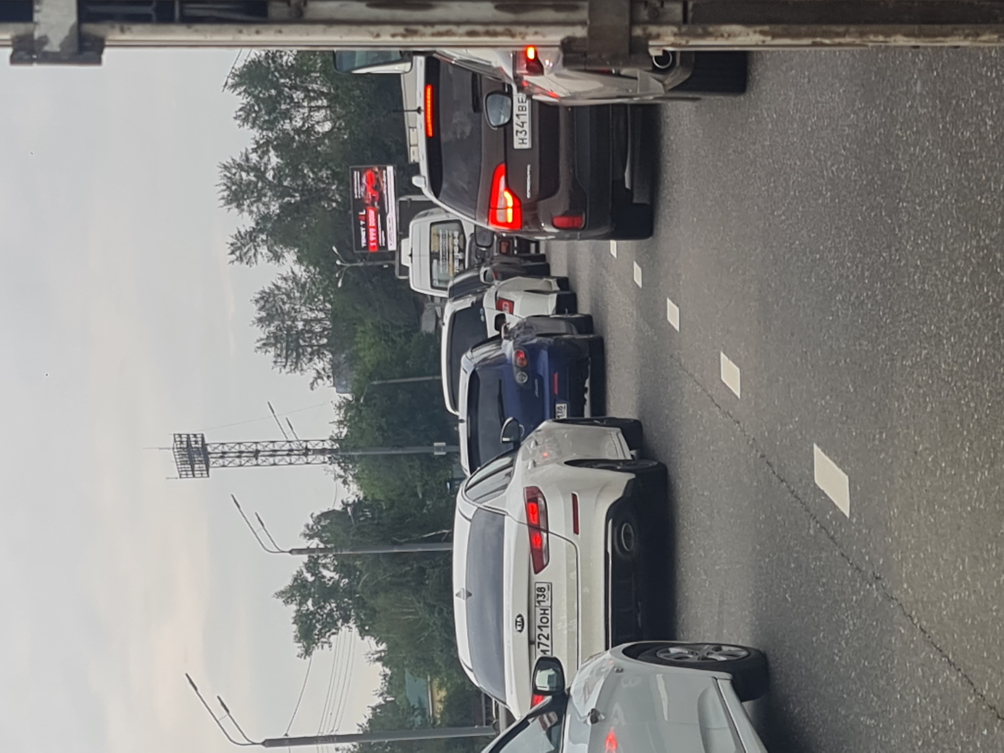 A traffic jam is visible on a cloudy day in Irkutsk, Russia, with numerous cars stopped on a multi-lane road. In the foreground, a white Kia Optima with a license plate reading "A721OH 138" is prominent. Behind it, a dark blue Mazda and a brown Kia Sorento with license plate "H341BE" are also visible. Other vehicles, including a white van and several more cars, are in the queue. Trees line the side of the road, and a tall lattice tower, possibly a communication tower, stands in the background. A large digital billboard displaying advertisements is also present. The road surface shows some cracks and is marked with white lane lines.