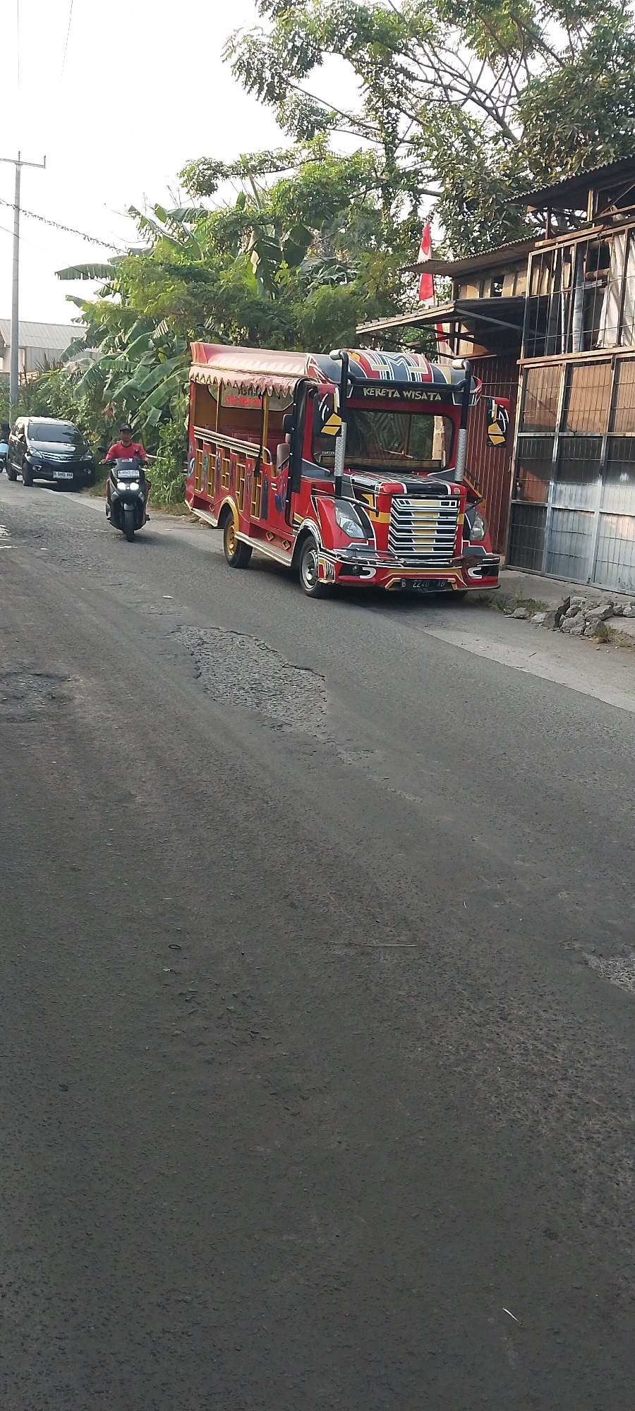 A bright red, open-sided tourist vehicle with a yellow and black patterned roof is parked on a paved road in Satriajaya, Indonesia. The vehicle has "KERETA WISATA" written on its upper front in yellow letters. To the left of the tourist vehicle, a dark-colored SUV is visible, and in front of it, a person wearing a red shirt rides a black motorcycle. The road is dark asphalt with visible patches of repair. On the right side of the road, a building with a corrugated metal roof and wooden walls is present, with some debris in front of it. Trees and banana plants line the left side of the road behind the vehicles. A utility pole is also visible in the background.
