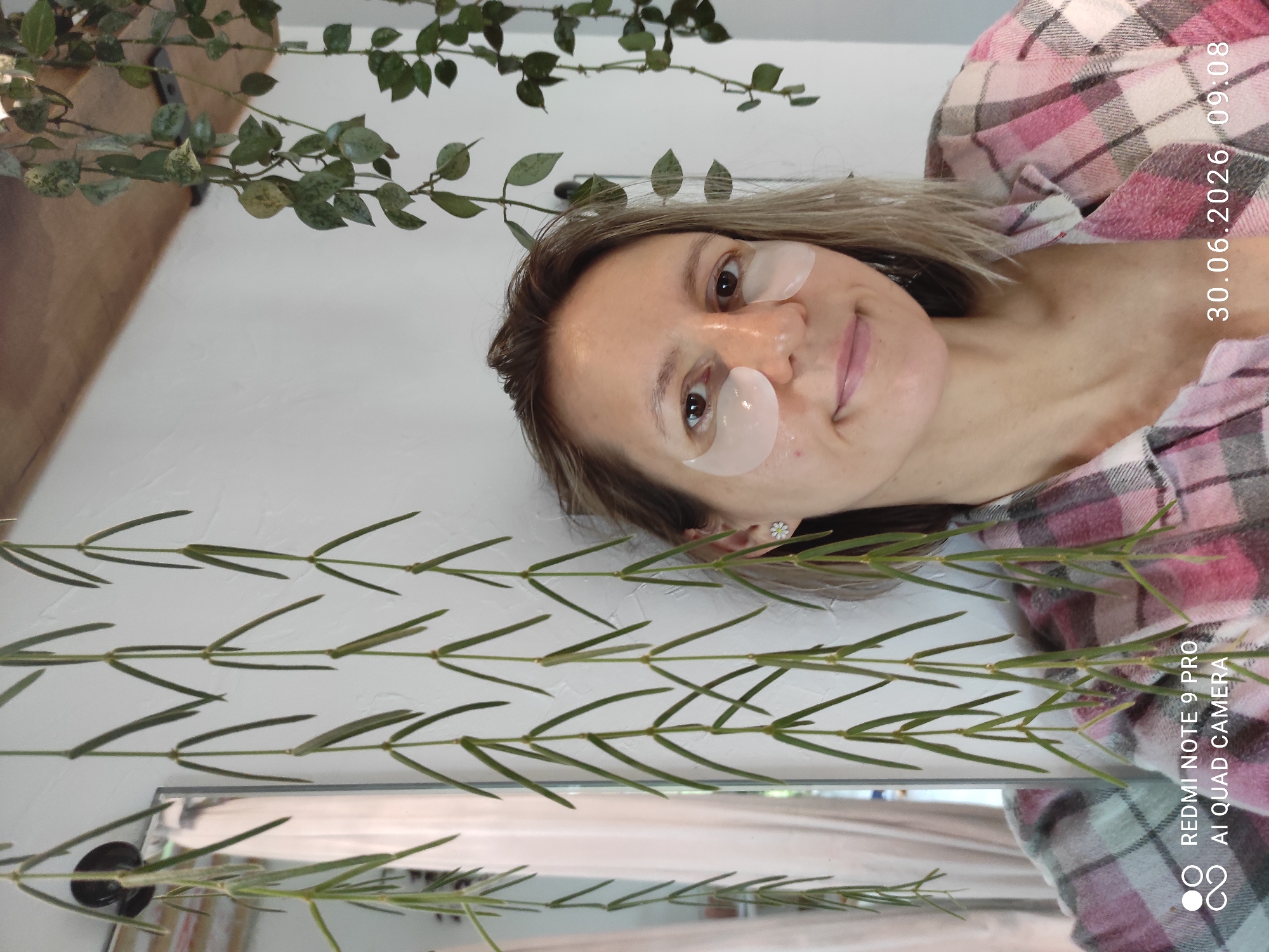 A woman is taking a selfie in front of a white wall. She is wearing eye patches and a pink and white plaid shirt. She has blonde hair and is smiling. There are plants hanging in the background, and a mirror is visible to the left of the woman. A timestamp in the lower right corner indicates the image was taken on June 30, 2026, at 09:08. The location "REDMI NOTE 9 PRO AI QUAD CAMERA" is displayed in the lower left corner.