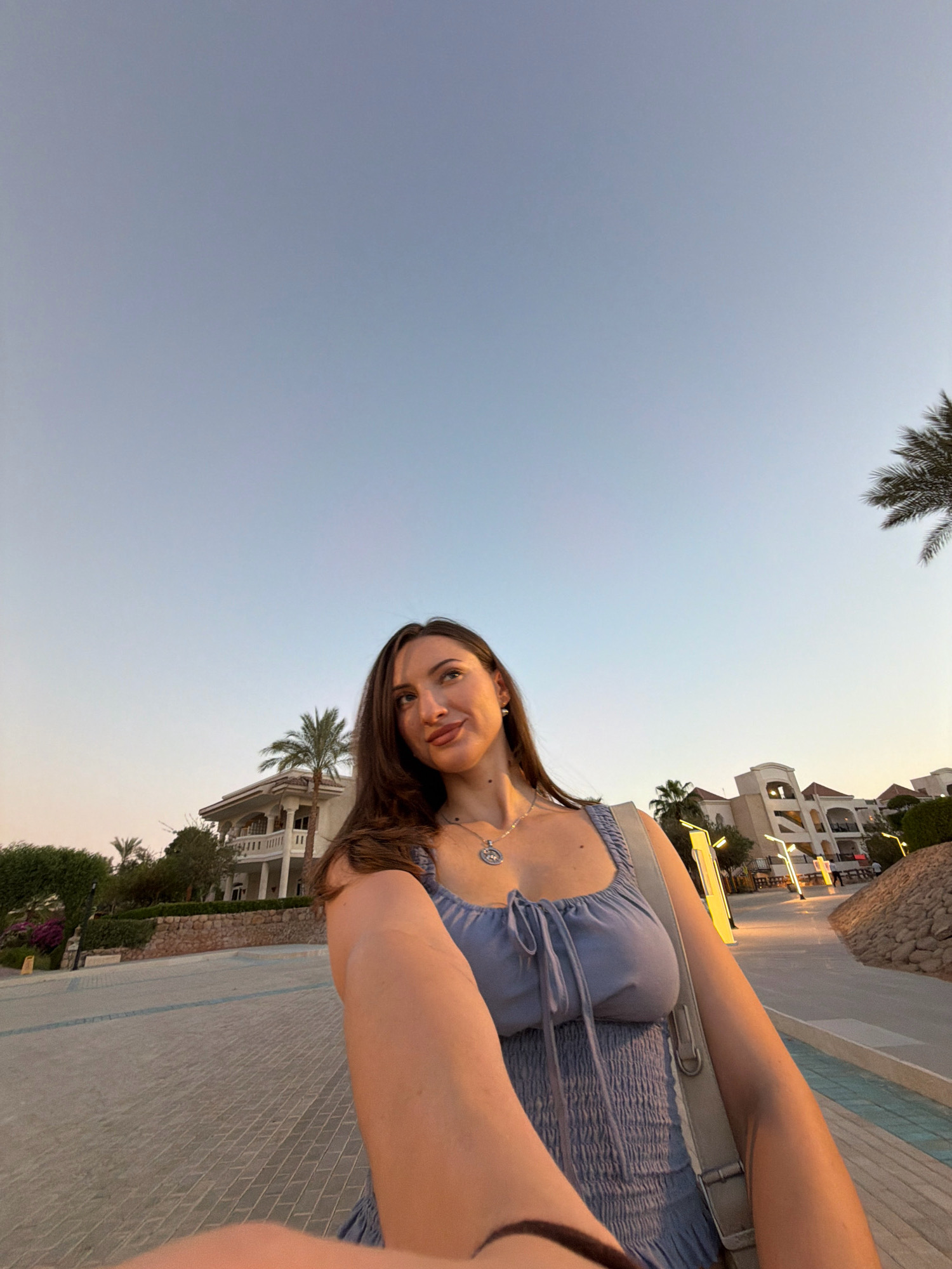 A young woman is captured in a selfie-style shot during what appears to be sunset in Sharm El Sheikh, Egypt. She is wearing a light blue, smocked sundress with a tie-front detail and a silver pendant necklace. Her long, dark hair is down, and she is looking upwards and to her right with a slight smile. Behind her, a modern, white building with balconies and palm trees is visible. The ground is paved with stone tiles. The sky is a clear, pale blue, suggesting the end of the day. The scene is set in an outdoor, likely resort or residential area, with streetlights illuminated in the background.