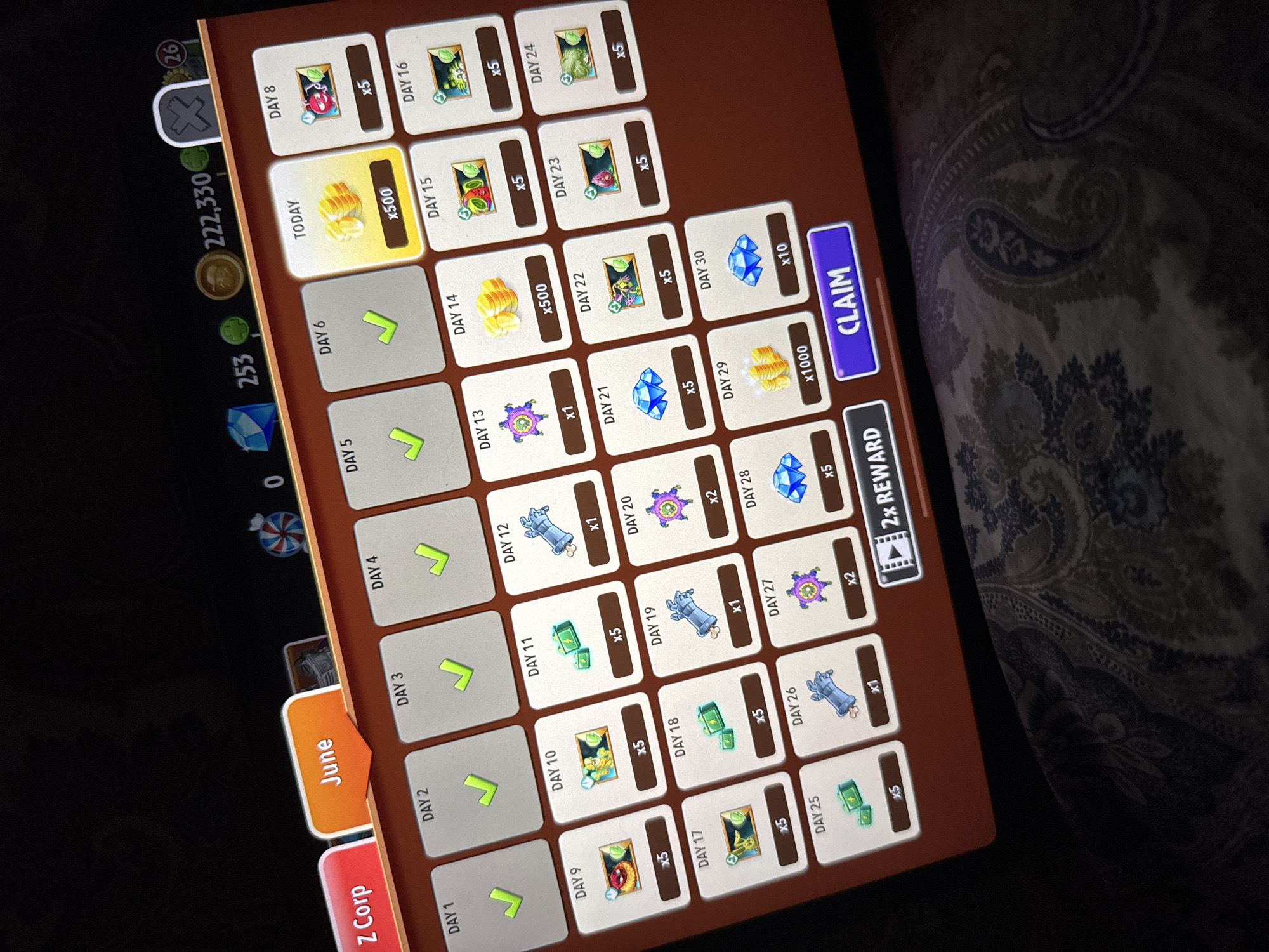 The image shows a digital screen displaying a calendar interface for a game, likely a mobile game, with a "June" tab selected. The interface is organized into daily reward slots, with some days marked with checkmarks, indicating claimed rewards. Various rewards are depicted, including coins, gems, and game items represented by icons. There is a prominent "CLAIM" button at the bottom right of the screen. The game's currency is also visible at the top, showing "222,330" coins and "253" gems. The background of the image is dark, with a portion of a patterned fabric visible in the lower half, suggesting the screen is placed on a surface. No specific location in Riga, Latvia is depicted or implied by the image content.