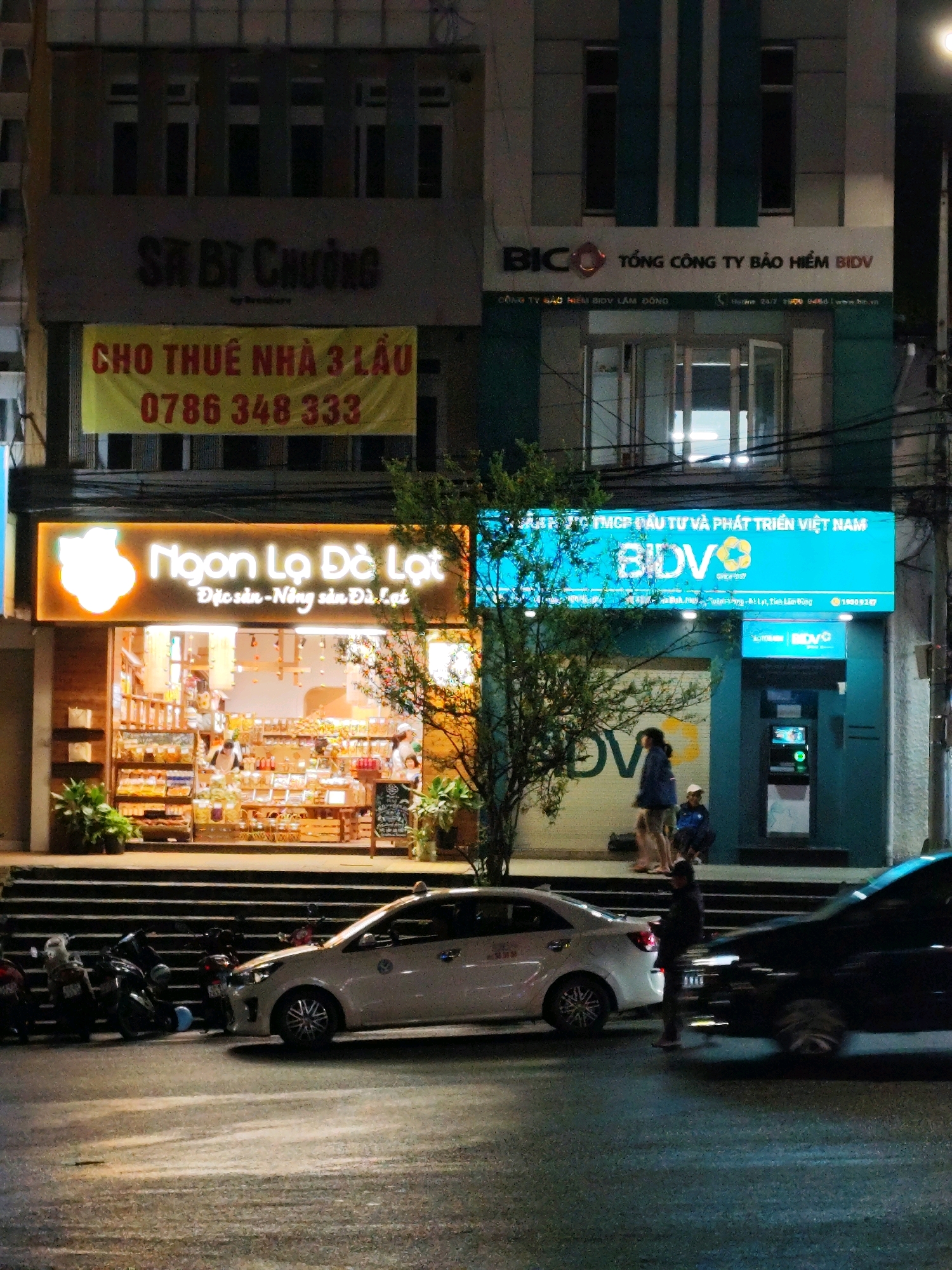 The image captures a nighttime street scene in Da Lat, Vietnam, featuring commercial establishments and street activity. On the left, a building displays a dark sign reading "SR BÀ CHƯỜNG" above a bright yellow banner with red Vietnamese text and the phone number "0786 348 333", indicating a property for rent. Below this, a warmly lit storefront with a neon sign that says "Ngon Lạ Đà Lạt" (Delicious Strange Da Lat) and "Đặc sản - Nông sản Đà Lạt" (Specialties - Da Lat Agricultural Products) is visible. This shop appears to sell local products, with shelves stocked with jars and bags.
To the right, a building with a prominent blue sign for "BIDV TỔNG CÔNG TY BẢO HIỂM" (BIDV General Insurance Corporation) is present. Further to the right, another blue BIDV sign is mounted above an ATM.
In the foreground, several motorcycles are parked on the left. A white car is parked in the middle of the street, and a darker vehicle is to its right, with its headlights illuminated. Several people are present: two individuals are standing near the BIDV building, and another person is standing near the white car. The street is wet, reflecting the lights. The overall ambiance is that of a bustling urban environment at night.