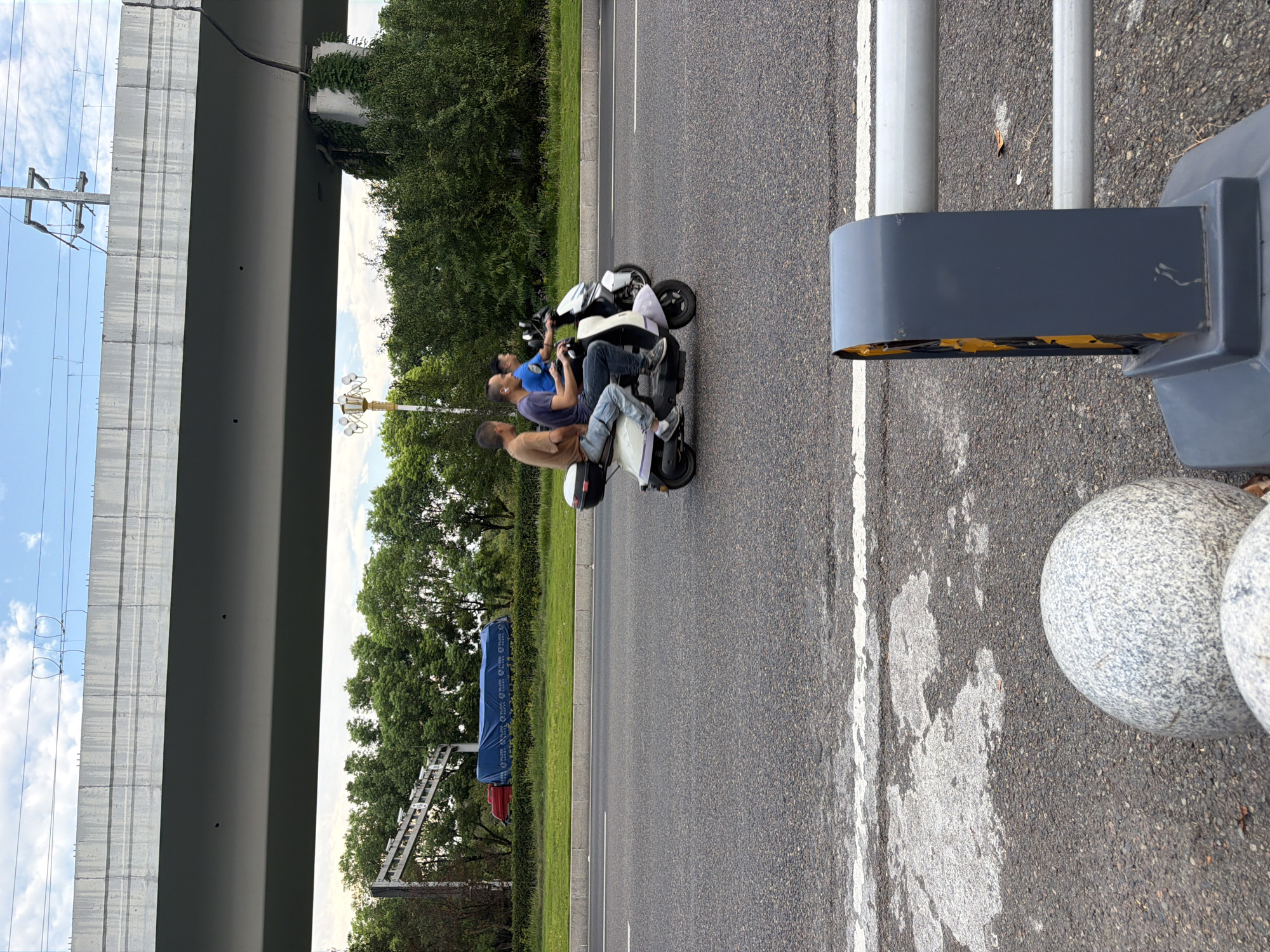 In Jiangjin, China, three individuals are captured on a scooter traveling along a multi-lane road, beneath an elevated overpass. The scooter, a white and light purple model with a black storage box, carries three people. The driver, wearing a blue shirt and jeans, operates the vehicle. Behind him, a man in a brown shirt and jeans sits as a passenger, and another man in a blue shirt and dark pants is positioned behind the driver. The road surface is asphalt with visible white lane markings. The environment includes lush green trees lining the roadside and a large concrete overpass overhead. Electrical wires and poles are also visible. In the foreground, a grey bollard with yellow reflective elements and a metal railing are present. The sky above is partly cloudy with a blue hue.