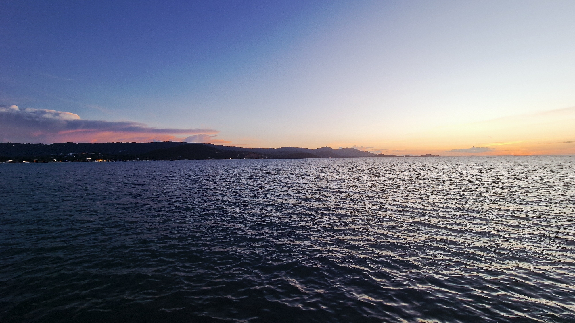 The image depicts a serene seascape at dusk, likely in the vicinity of Baan Plai Laem, Thailand, given the characteristic hilly coastline and the tranquil waters. The sky transitions from a deep indigo at the zenith to softer blues and then to a warm gradient of pale yellow and orange near the horizon, indicative of the sunset. Wispy clouds, tinged with pink and orange, are visible on the left side of the sky.
The water in the foreground is a dark, deep blue, reflecting the colors of the sky. Small, consistent ripples across the surface suggest a gentle breeze. The middle ground is occupied by the sea, with a dark, silhouetted coastline forming the background. The coastline is characterized by rolling hills, and scattered lights indicate human settlement, possibly a village or resort area along the shore. The overall impression is one of tranquility and natural beauty at the close of day. There are no visible people, objects, or actions that indicate any activity beyond the natural environment.