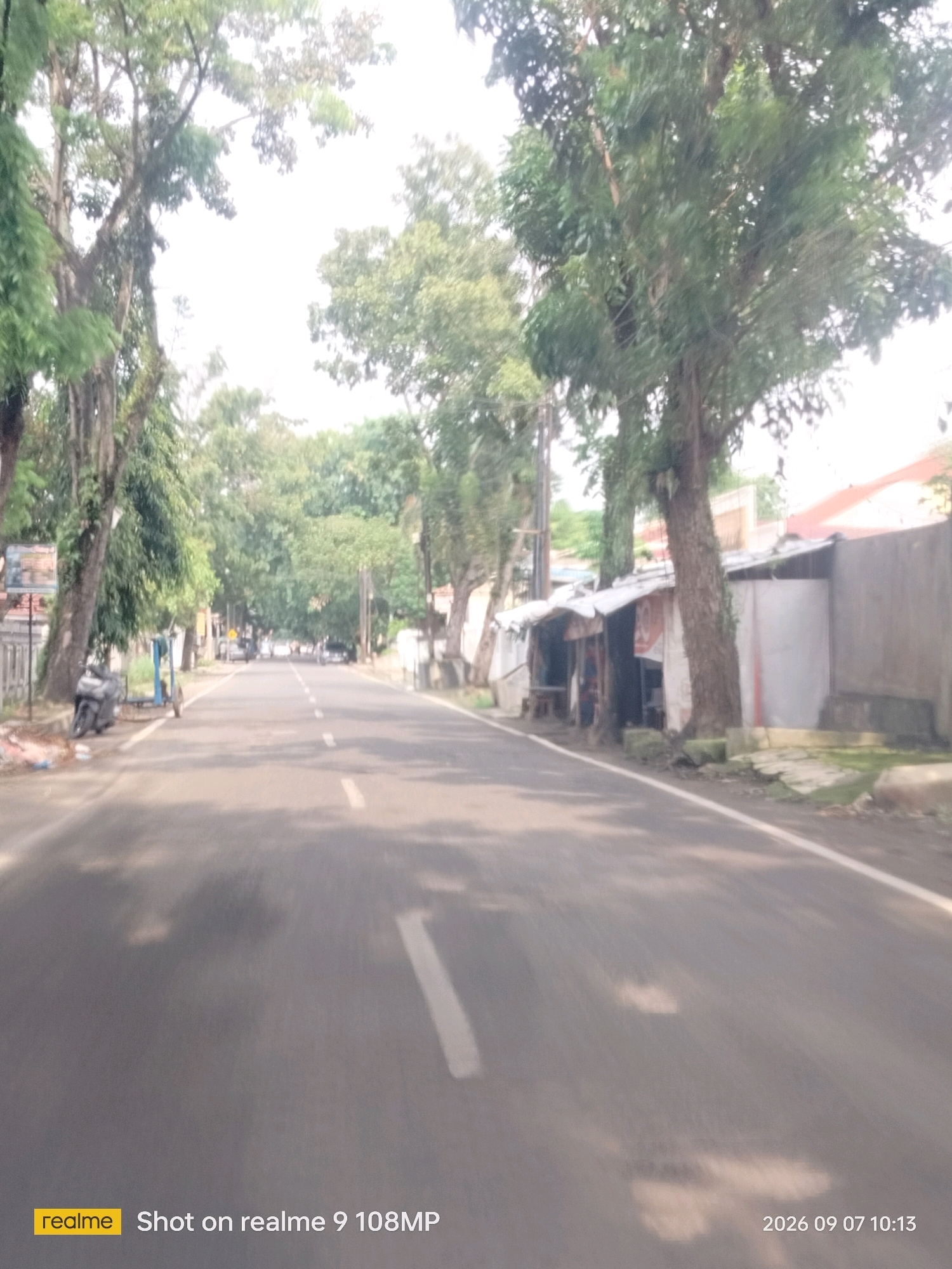 The image captures a street scene in the city of Medan, Indonesia, on September 7, 2026, at 10:13 AM. The road is paved and appears to be a two-lane street with a white line marking the center. On the left side of the road, there are several trees with dense foliage lining the sidewalk. A motorcycle and a cart are parked on the side of the road. A small structure, possibly a shop or stall, is visible on the right side of the road, constructed with corrugated metal and wood, and appears to be built around a large tree. The background shows more trees and buildings, suggesting a moderately developed urban or suburban area. The sky is overcast or hazy, creating a bright, diffused light. The image is a candid shot, likely taken from a moving vehicle, as indicated by the motion blur in the foreground. The "realme Shot on realme 9 108MP" watermark is present in the lower left corner.