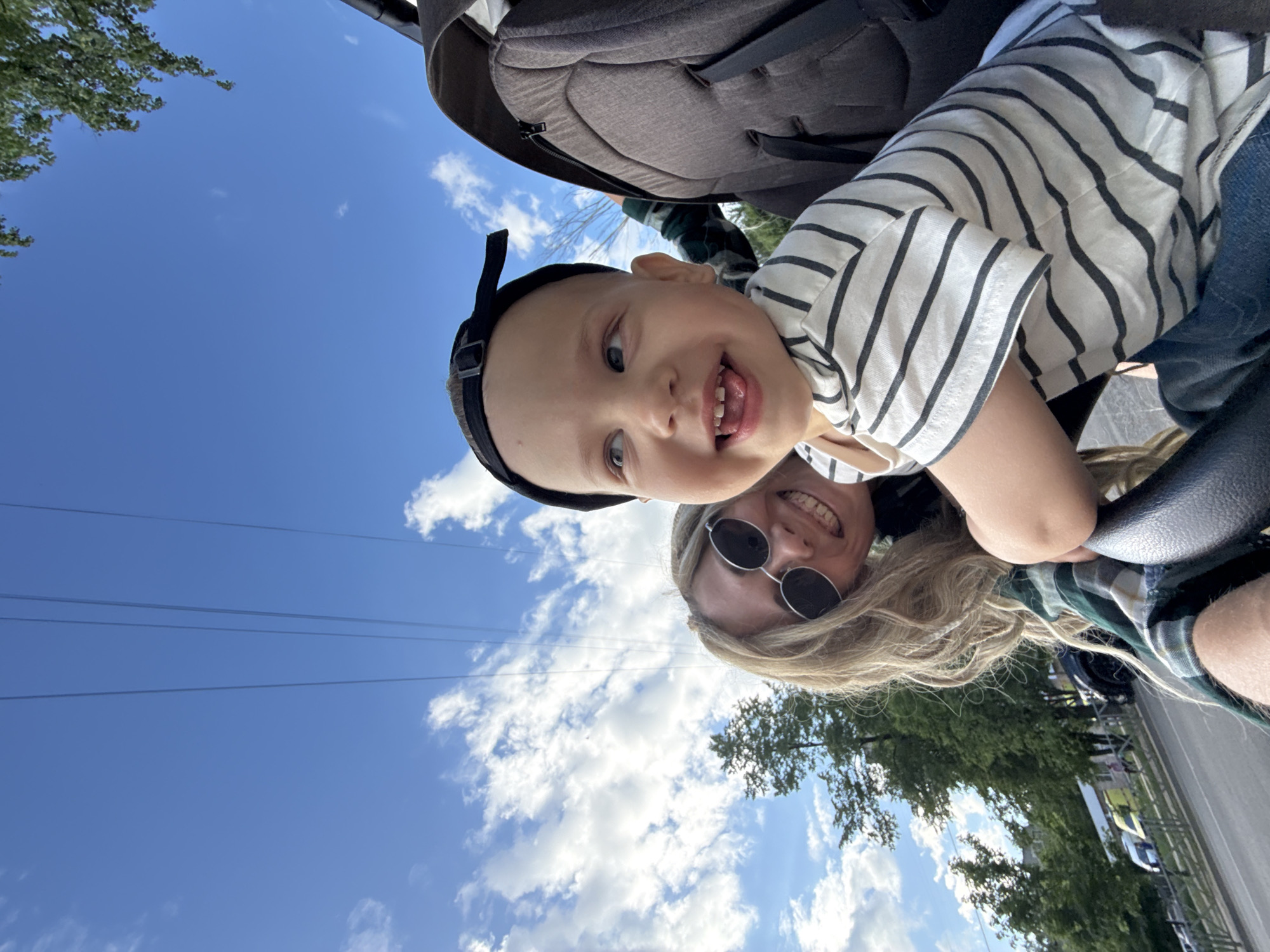 The image shows a selfie taken outdoors on a sunny day, likely in the city of Rusanovo, Russia. The camera angle is tilted. A young child, possibly a toddler, is in the upper part of the frame, wearing a striped shirt and a black baseball cap. The child is sticking their tongue out and smiling. Below the child is an adult, likely female, wearing sunglasses and a green and white plaid shirt. The adult is smiling.
Above the child, part of a baby stroller is visible. In the background, a bright blue sky with scattered white clouds is seen, along with power lines and trees which can be assumed to be a part of Rusanovo based on the data provided. A street and parked cars are visible near the bottom of the frame.