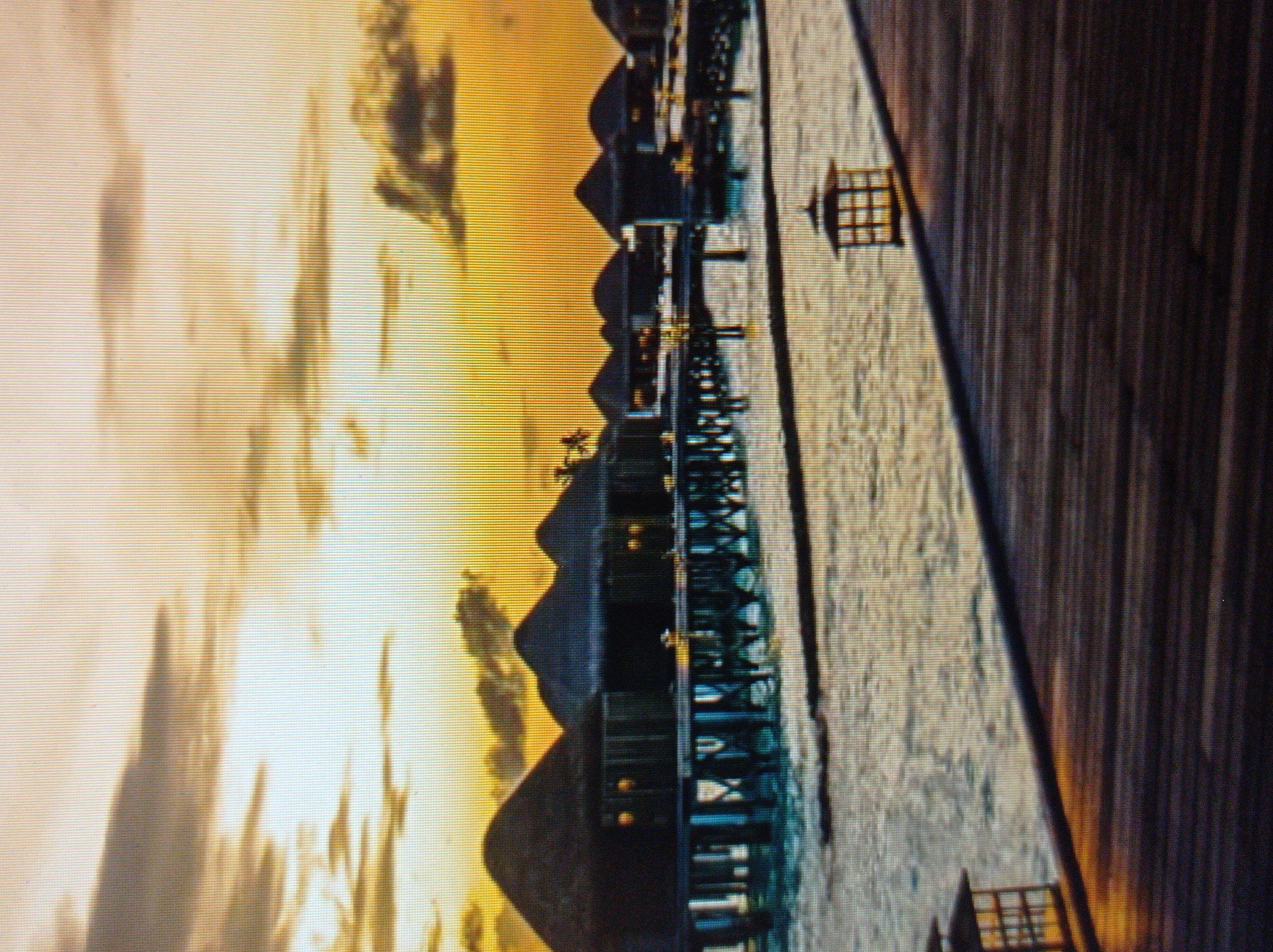 This is a photograph of overwater bungalows at sunset. The sky is a gradient of yellow and orange, with scattered clouds. The bungalows are dark and silhouetted against the sky, with thatched roofs and stilts. They are situated over calm water, and a wooden boardwalk leads towards the foreground of the image. There is a lantern on the boardwalk. The scene appears to be a resort. There is no indication of the city of Mukachevo, Ukraine, in this image.