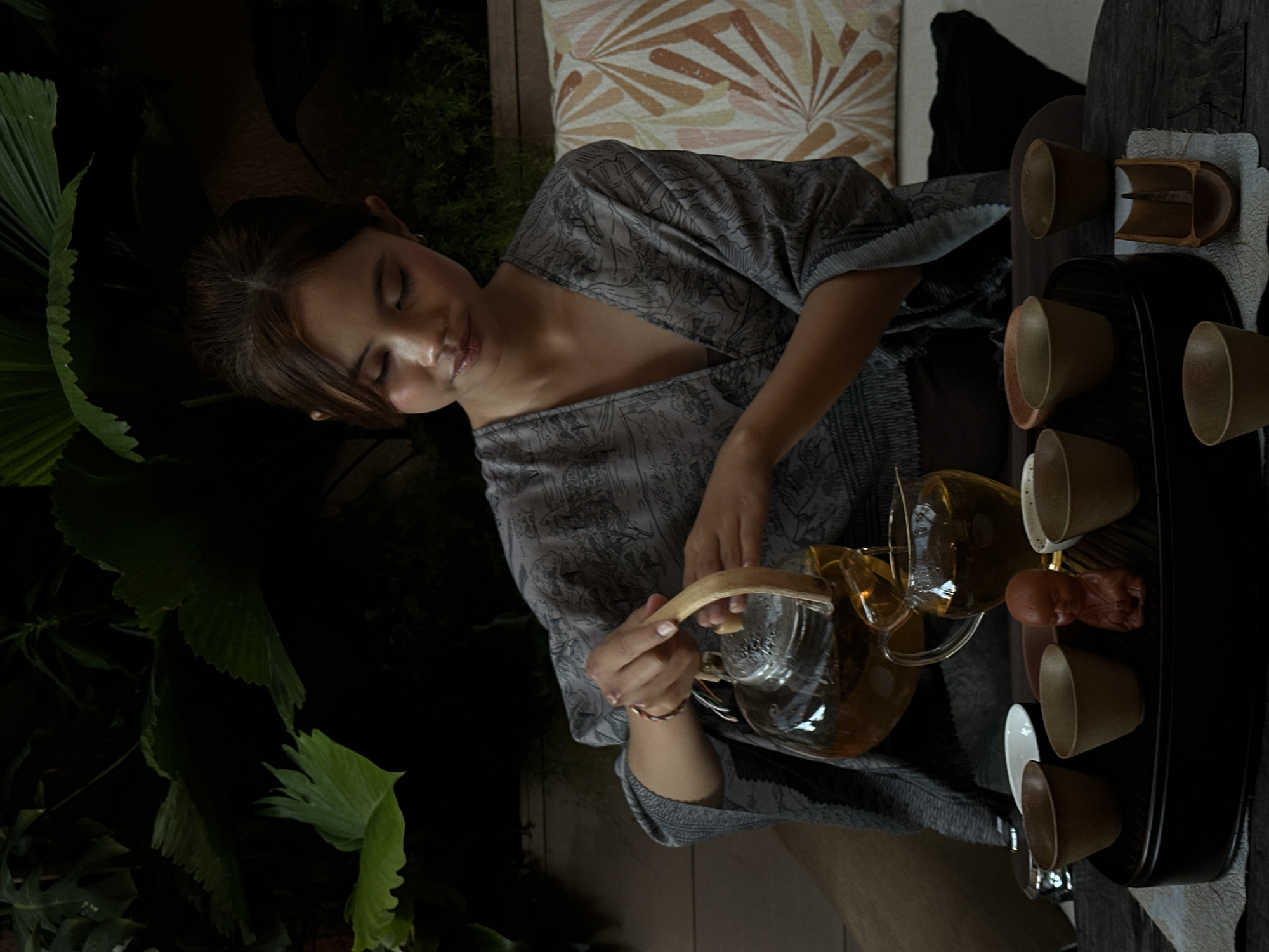 A woman is shown holding a clear teapot filled with a yellowish liquid, likely tea, pouring it. She is seated outside near some greenery. A black tray beside her holds several small cups. The location is Pecatu, Indonesia.