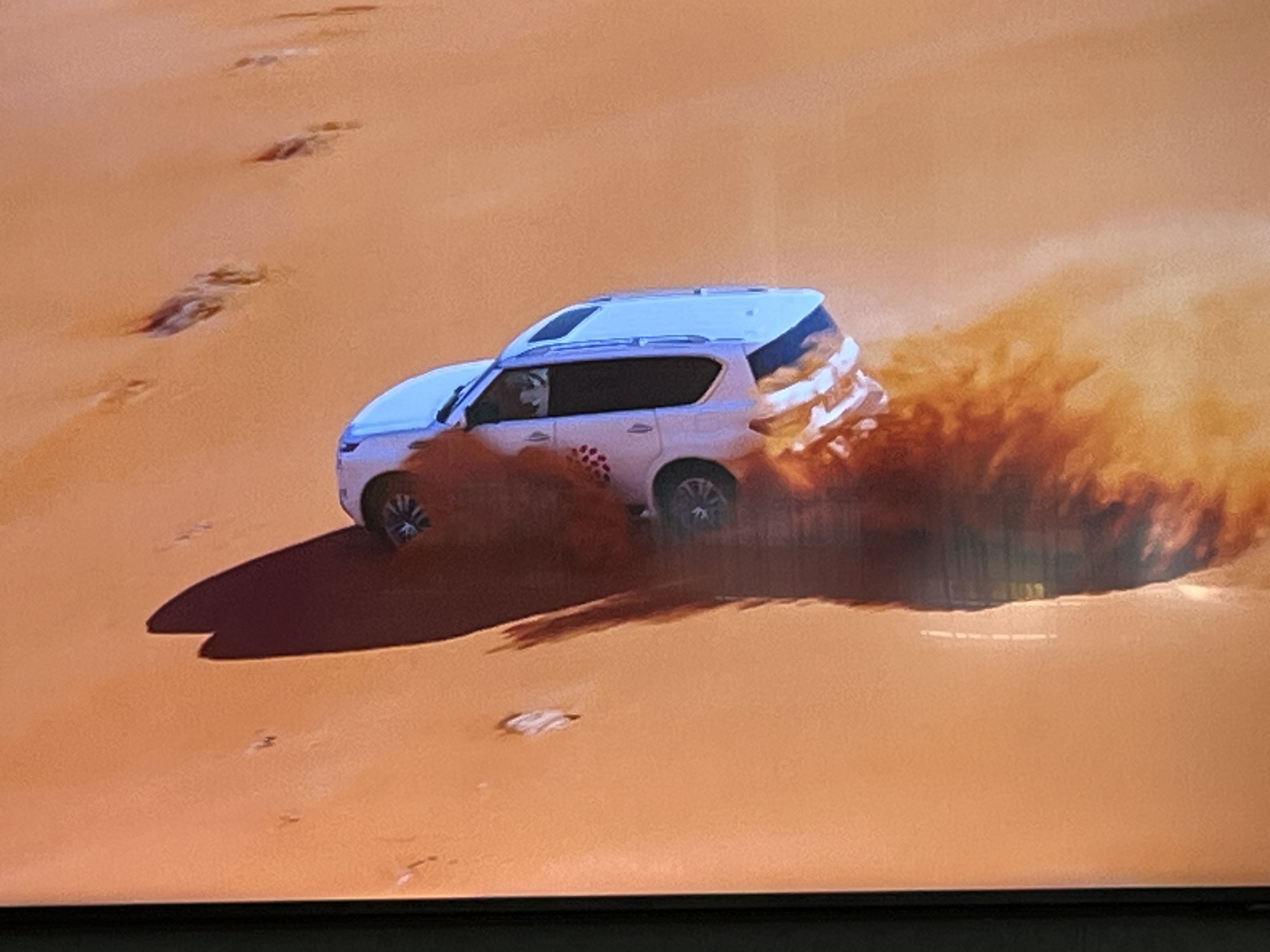 The image depicts a white SUV driving on a sand dune, kicking up a large amount of sand. The vehicle is angled, suggesting it is in the process of a turn or maneuver. The environment is a desert landscape with a clear, orange-tinted sky. The sand is smooth and undulating, with some tire tracks visible in the foreground and mid-ground. There are no people visible in the image. The scene does not provide any information or context to suggest a location in Kharkiv, Ukraine.