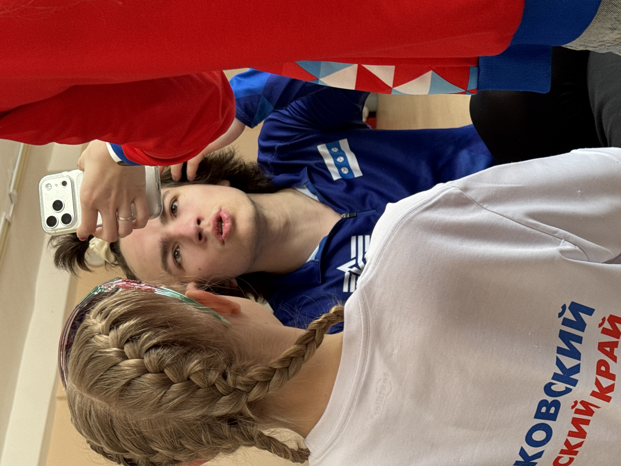 A photo shows three people, presumably taking a selfie. A person in a red sweater holds a smartphone. The smartphone is pointed down toward a second person wearing a blue Adidas zip-up track jacket with a white emblem that has three balls. The third person wears a white T-shirt with the words "Kovskiy Kray" written in blue and red. The city of Berezniki, Russia is not visible.