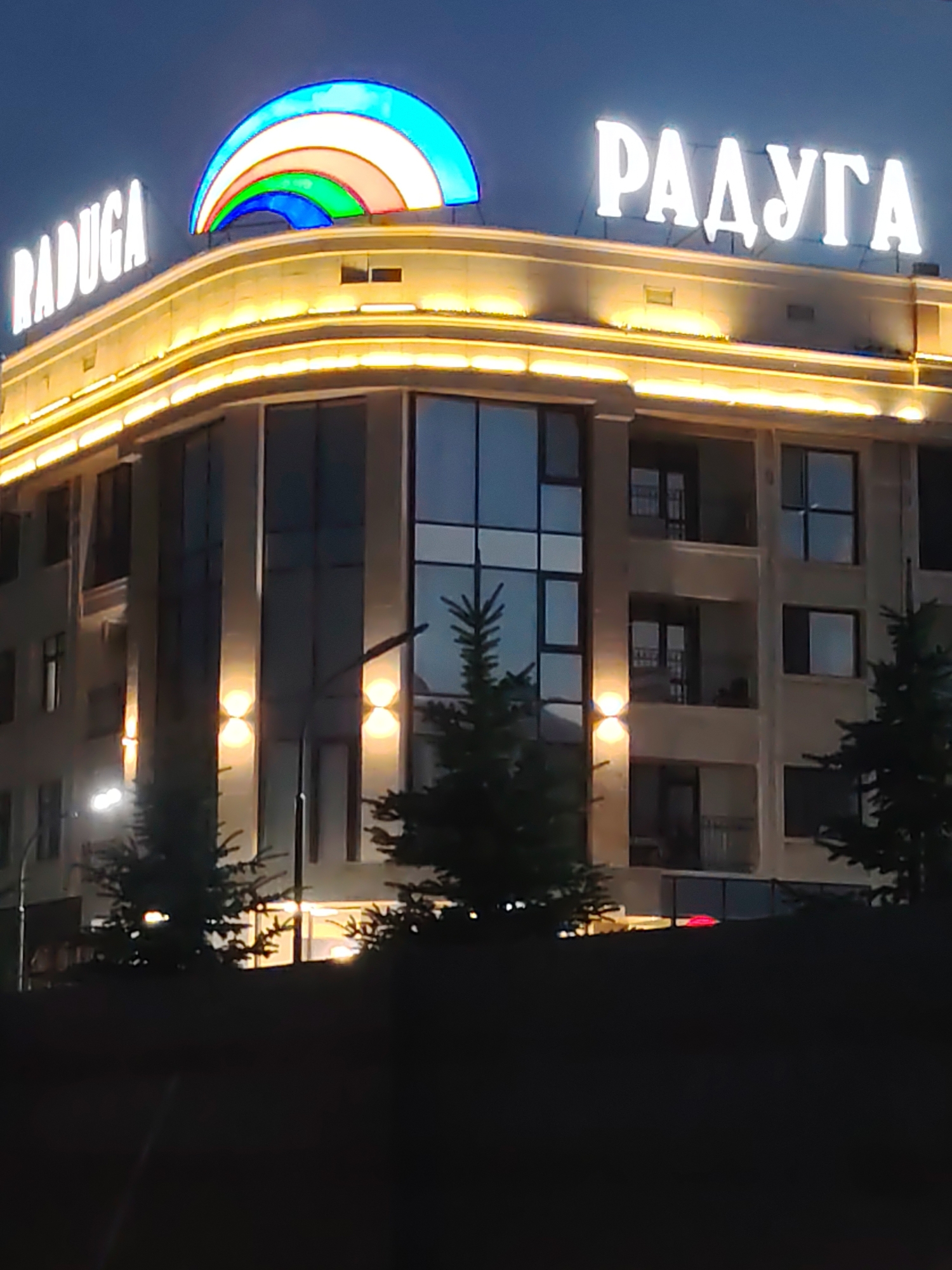 The image shows the "RADUGA" hotel in Sary-Oy, Kyrgyzstan, at night. The hotel has a prominent illuminated rainbow sign on its rooftop and the word "RADUGA" in illuminated white letters. The building is modern with multiple floors featuring large windows and balconies. Exterior lights illuminate the facade and accent the architectural details. Several evergreen trees are visible in the foreground, partially obscuring the lower levels of the building. The sky is dark, indicating nighttime. No people are clearly visible in the image.