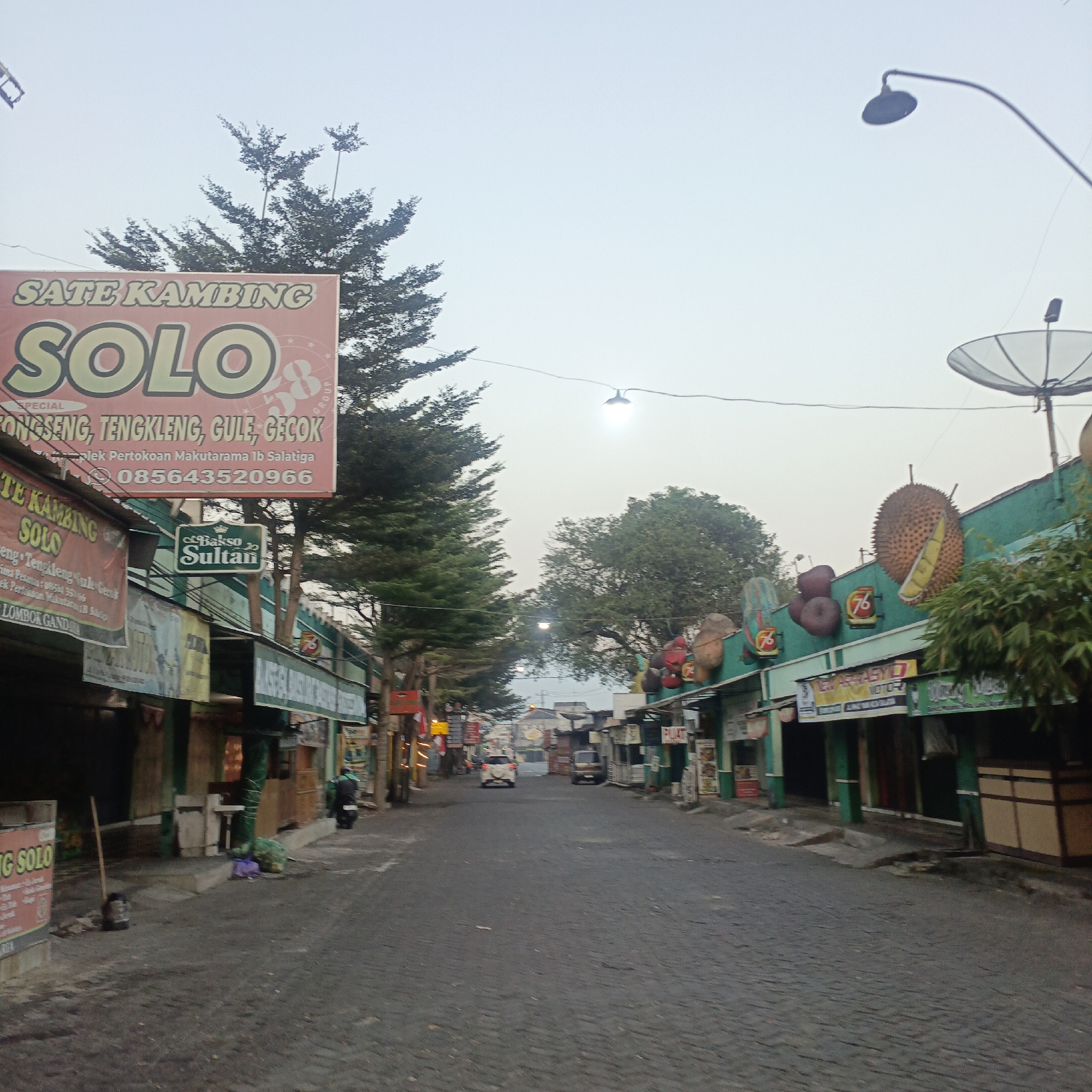 The image shows a street scene in Salatiga, Indonesia, with a focus on food stalls. The sky is overcast, suggesting either early morning or late afternoon. On the left side of the street, a prominent sign reads "SATE KAMBING SOLO", indicating a goat satay restaurant. Below this, another sign advertises "GULE, GECÓK, TENGKLENG". Further down the street, a smaller sign reads "Bakso Sultan", denoting a meatball restaurant. The buildings lining the street appear to be small shops or food stalls, many of them with green facades. Large, inflated durian fruit models are visible on top of one of the buildings, suggesting it might be a fruit-themed establishment or a restaurant specializing in durian-based dishes. A satellite dish is mounted on a pole above the durian decorations. The street itself is paved with dark cobblestones. A few vehicles, including a white car and a motorcycle, are present on the street, and a person in dark clothing is visible on the motorcycle. In the foreground, a broom and a trash bin are placed near a stall, indicating ongoing activity or preparation. The overall impression is a vibrant street market or dining area, typical of Indonesian culinary hubs.