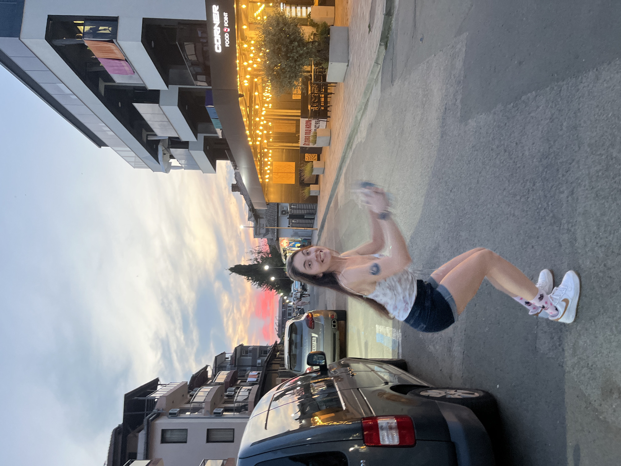 A young woman with long brown hair is captured mid-squat on a street in Lozenets, Bulgaria. She is wearing a white tank top with a floral pattern, denim shorts, and white sneakers with pink socks. Her arms are extended forward, and she appears to be holding something in her hands. Her facial expression is one of excitement or surprise, with a wide smile and open mouth.
The setting is a street with buildings on either side. To the left, a dark gray van is parked. Across the street, a modern building with multiple balconies and a lit sign for "CORNER FOOD & POINT" is visible, with strings of lights illuminating the outdoor seating area. Other buildings, trees, and streetlights are also present in the background. The sky displays a colorful sunset with hues of pink, orange, and blue. The overall atmosphere suggests a casual evening in a coastal town.